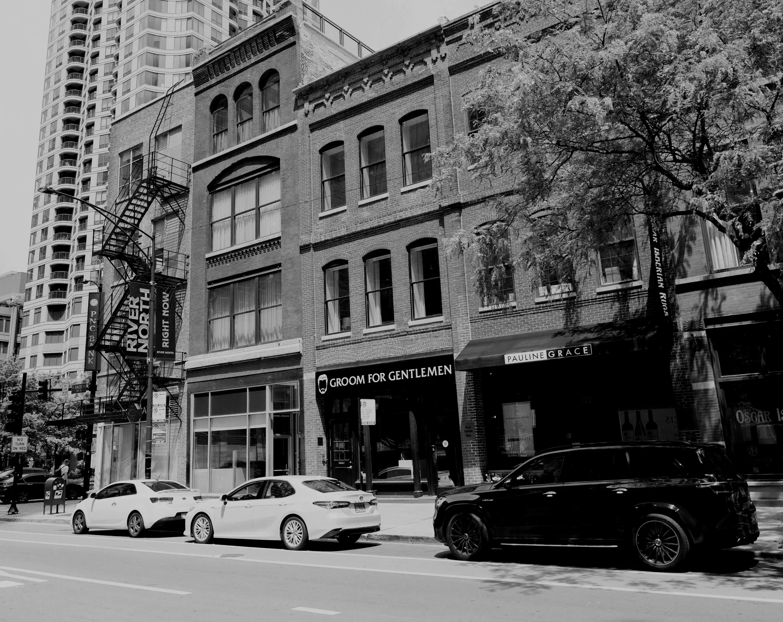 Black and white photograph of city street with modern high-rise buildings, historic brick building, parked cars, trees, and street signs.