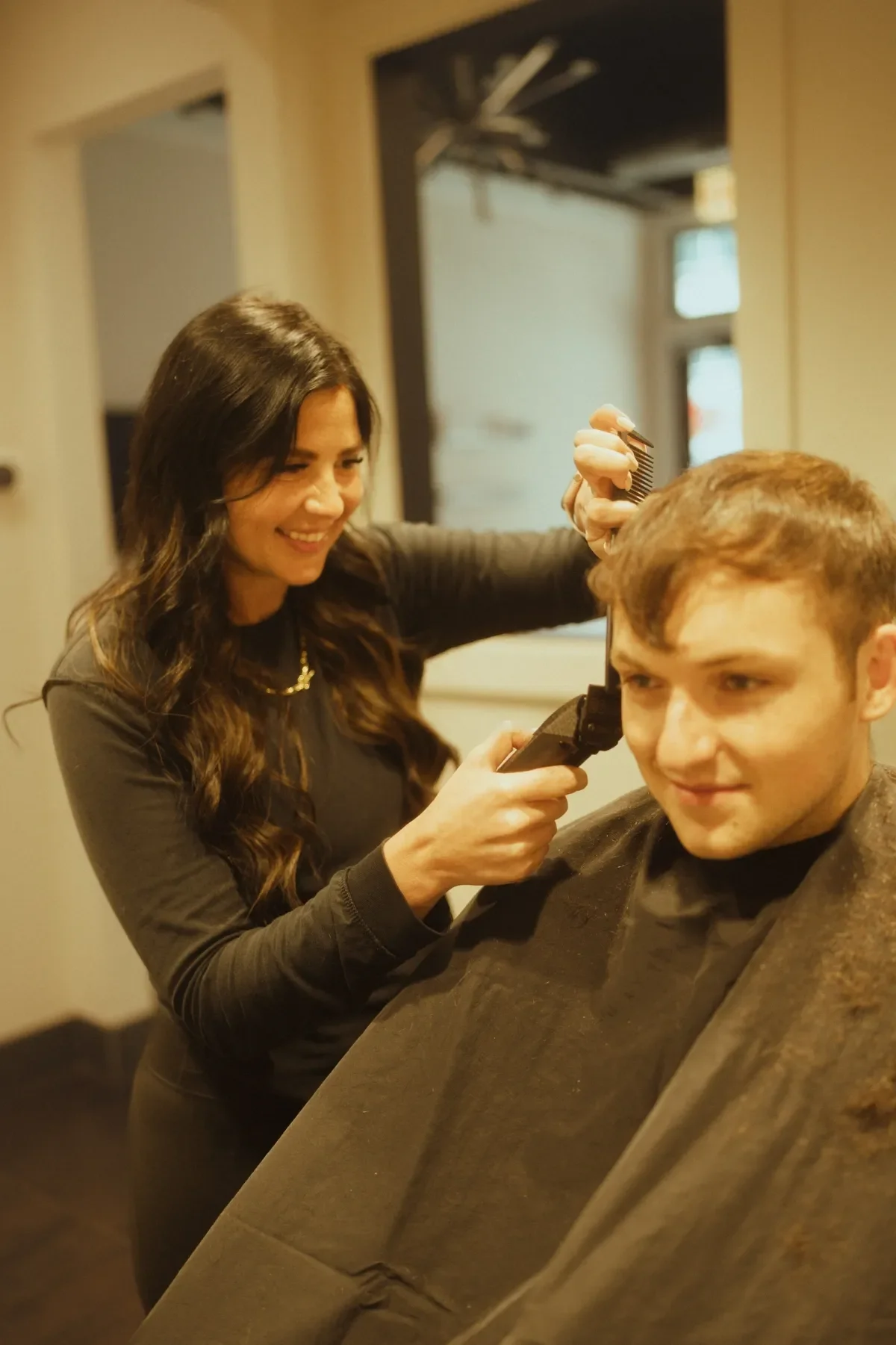 A woman cutting a young man's hair in a salon. The woman is smiling and holding hair cutting scissors, while the young man is sitting on a salon chair covered with a brown cape.