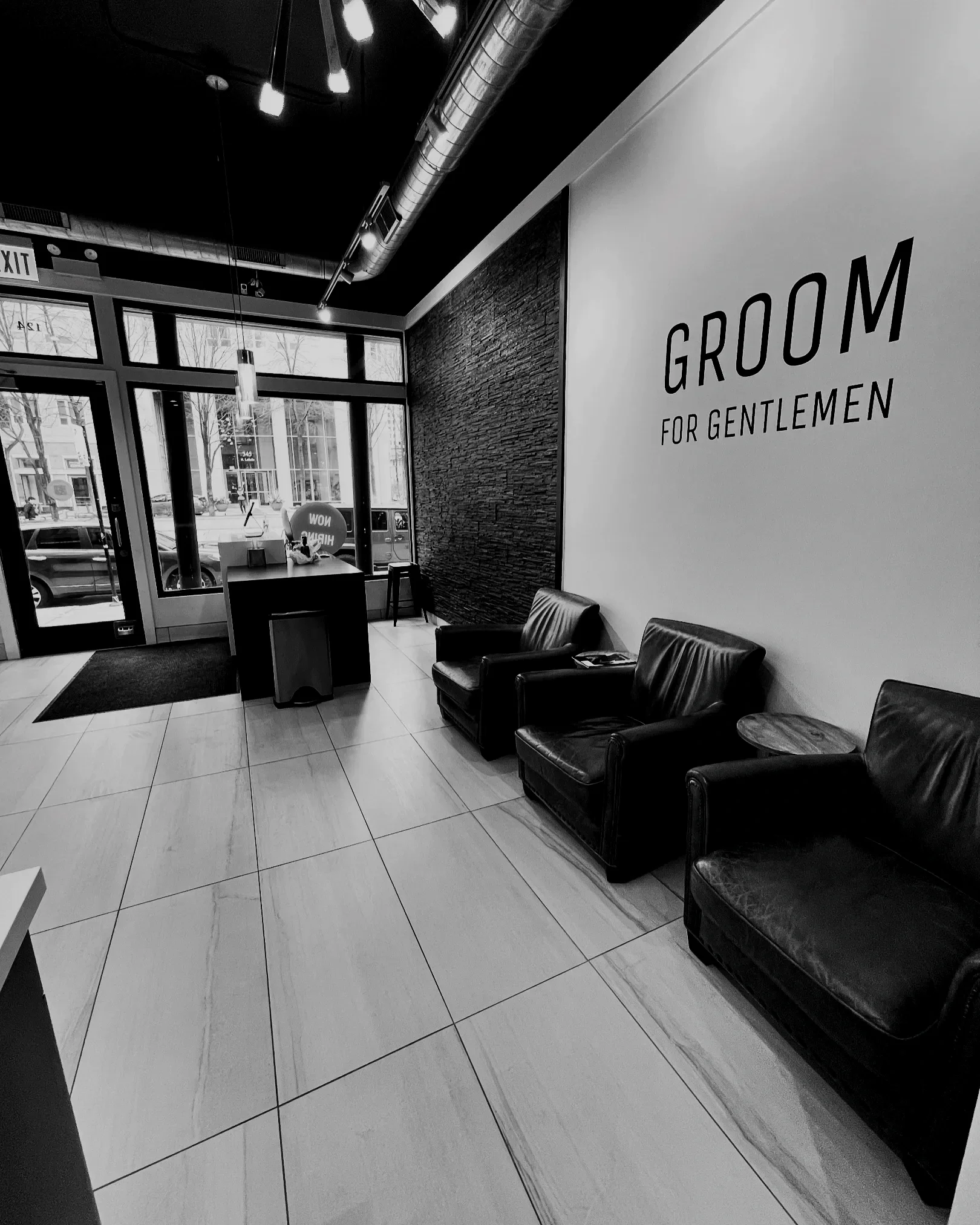 Interior of a salon with a sign on the wall reading 'GROOM FOR GENTLEMEN', black leather chairs, a small table, tiled floor, large windows, and visible ceiling ductwork and lighting.