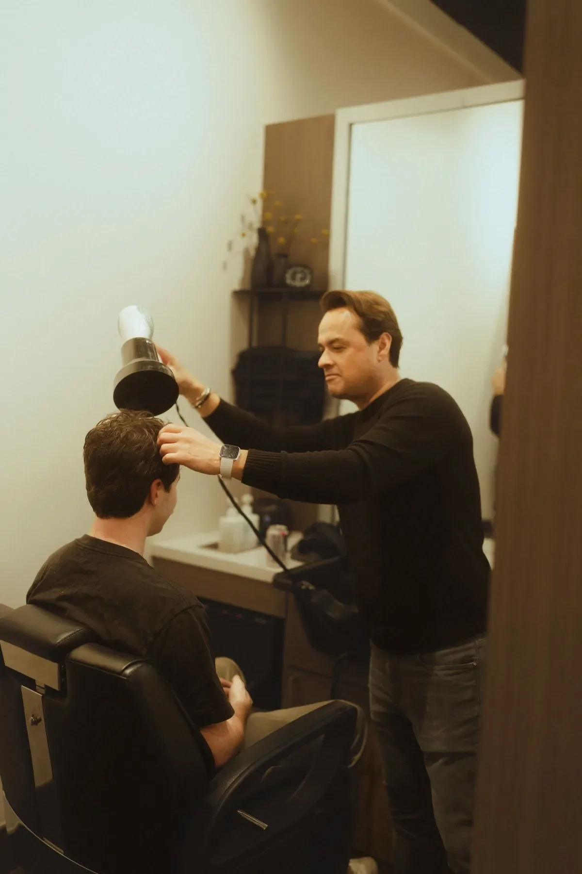 A hairstylist drying a man's hair with a blow dryer while he sits in a salon chair. The stylist is focused on his work, and the salon has a mirror, a counter with hair products, and decor in the background.