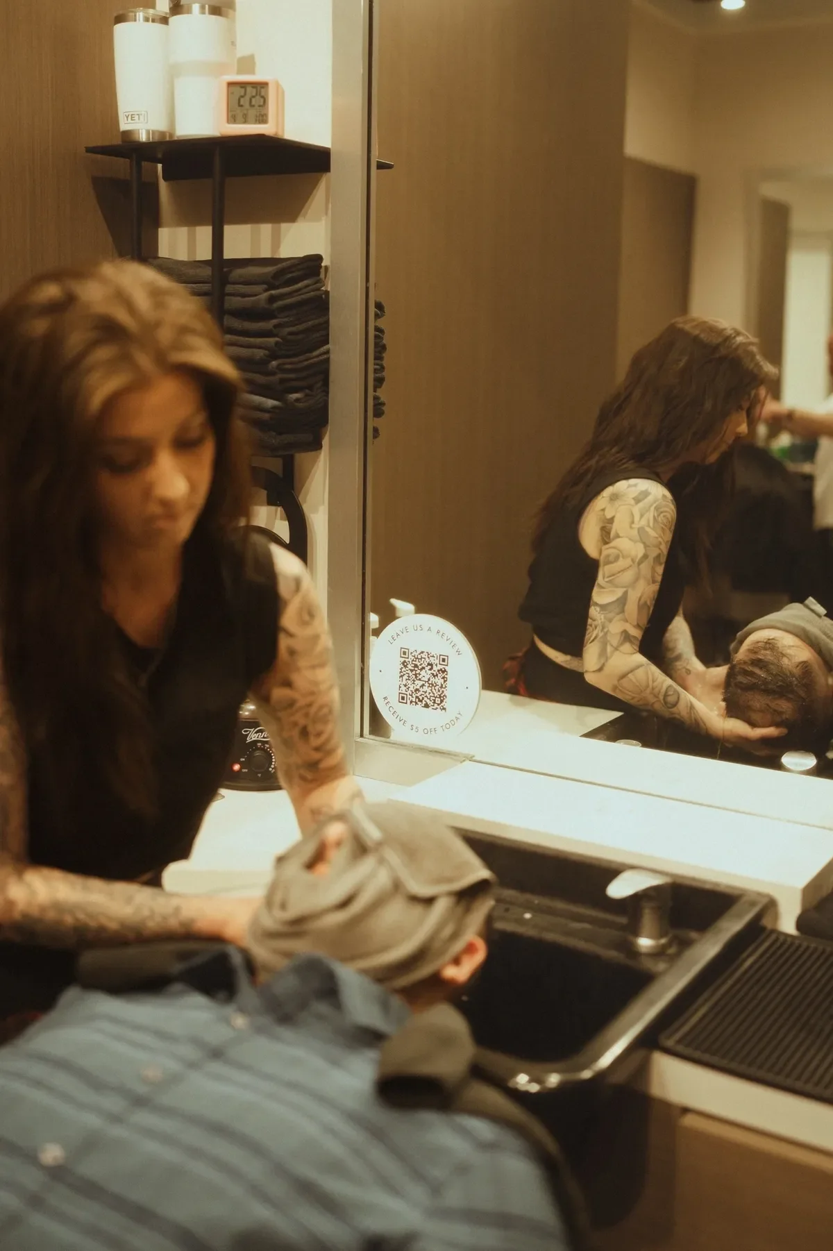 A tattooed woman at a salon station, holding a client's head, reflected in a mirror, with various towels and a QR code sticker on the mirror.