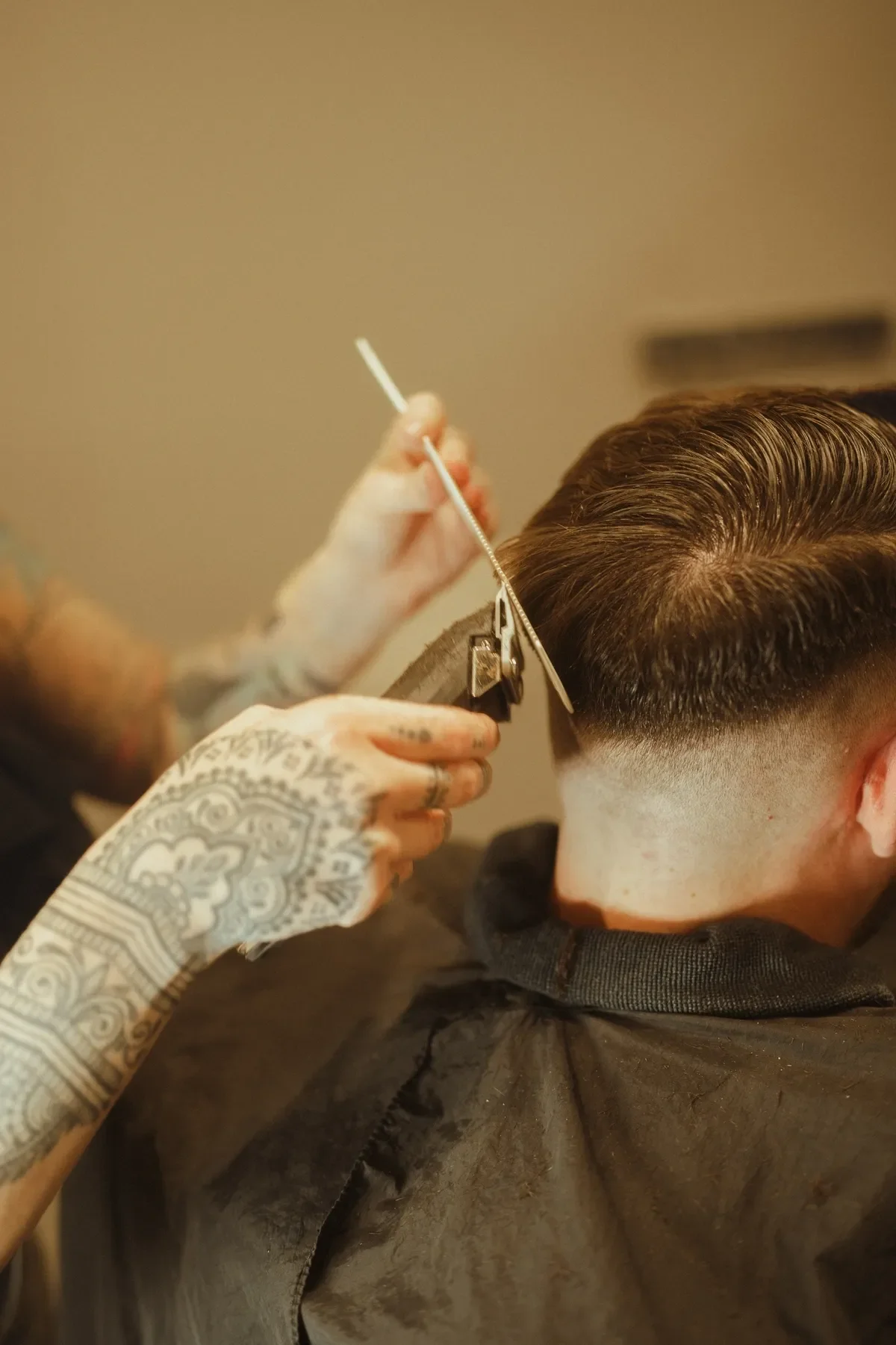 A person getting a haircut with a close-up of the barber's tattooed hand holding clippers, trimming the hair of a young man with a slicked-back hairstyle.