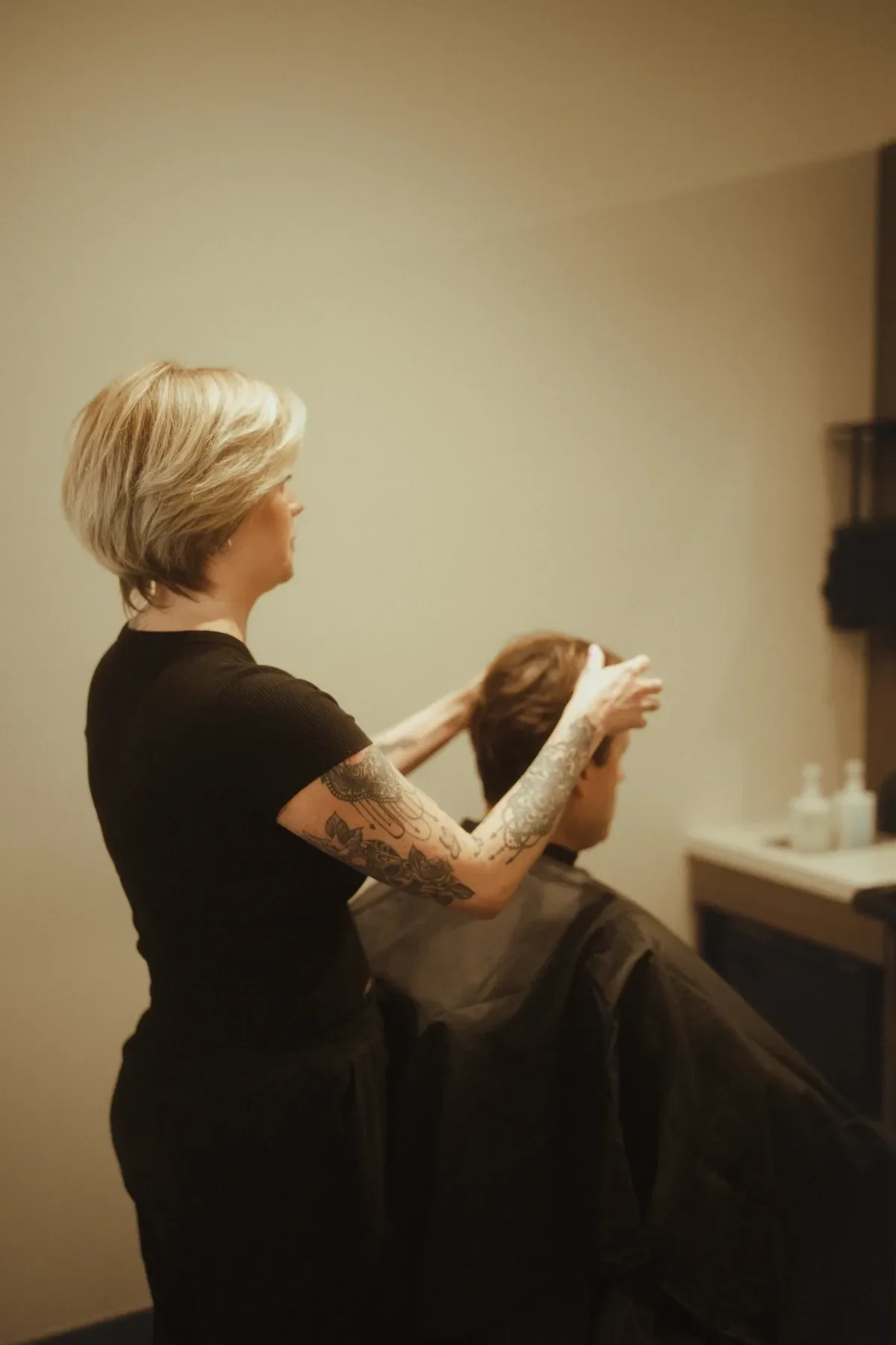A hairstylist with tattoos on her arm is styling a client's hair in a salon. The client is sitting in a chair with a black cape, and the background is a plain wall with some salon products on a counter.