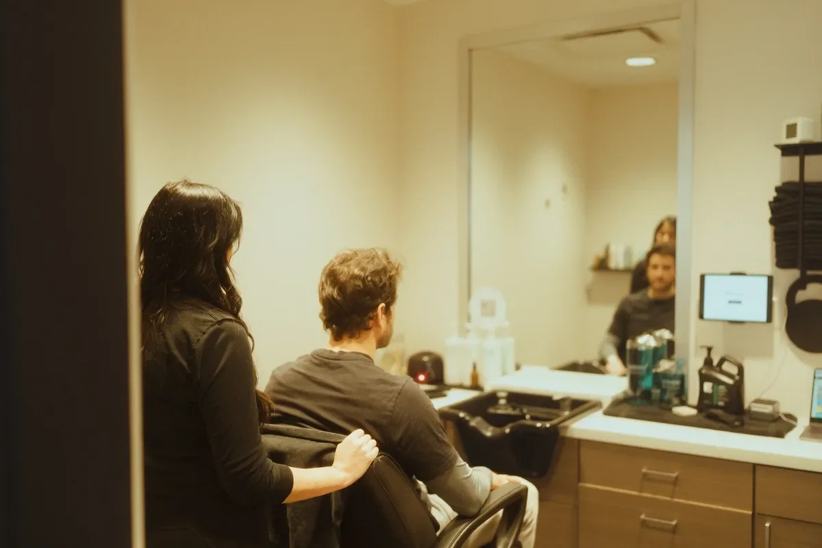A man and a woman sitting in a salon, facing a large mirror, with the woman standing behind the man, touching his shoulder, while the stylist and another client are seen in the mirror.