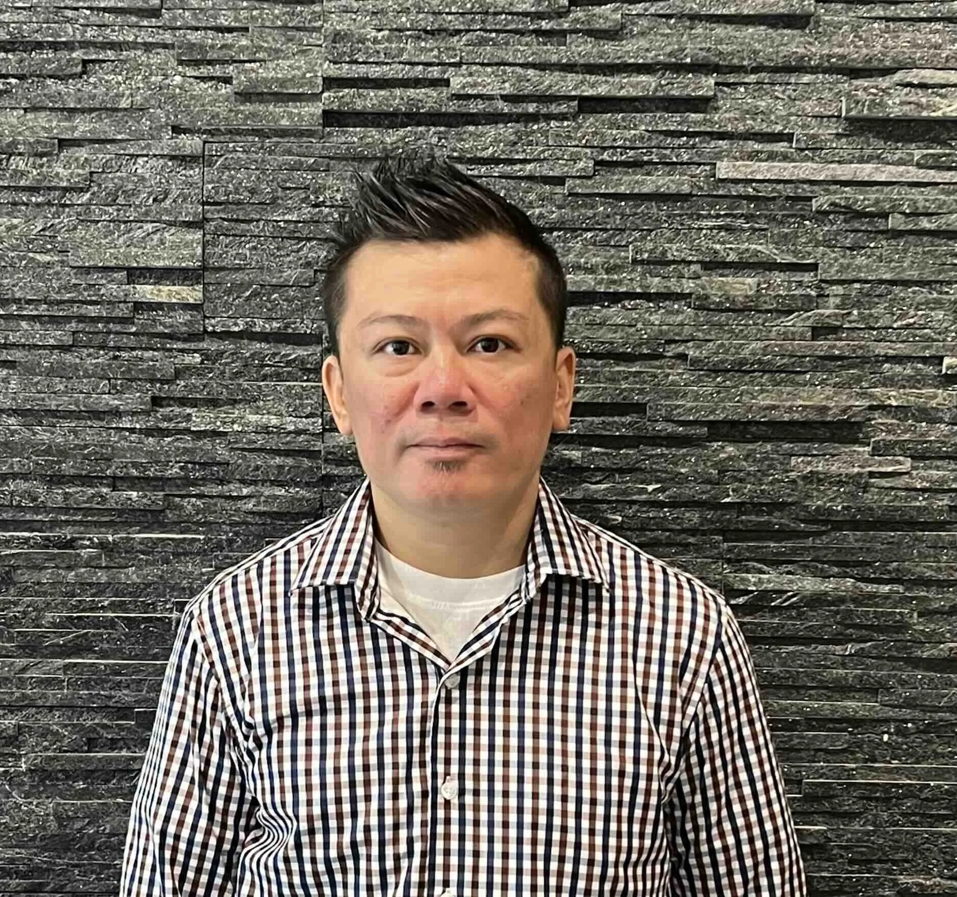 A man with short dark hair and a clean-shaven face wearing a checkered shirt white, black, and red with a white undershirt, standing in front of a textured stone wall.