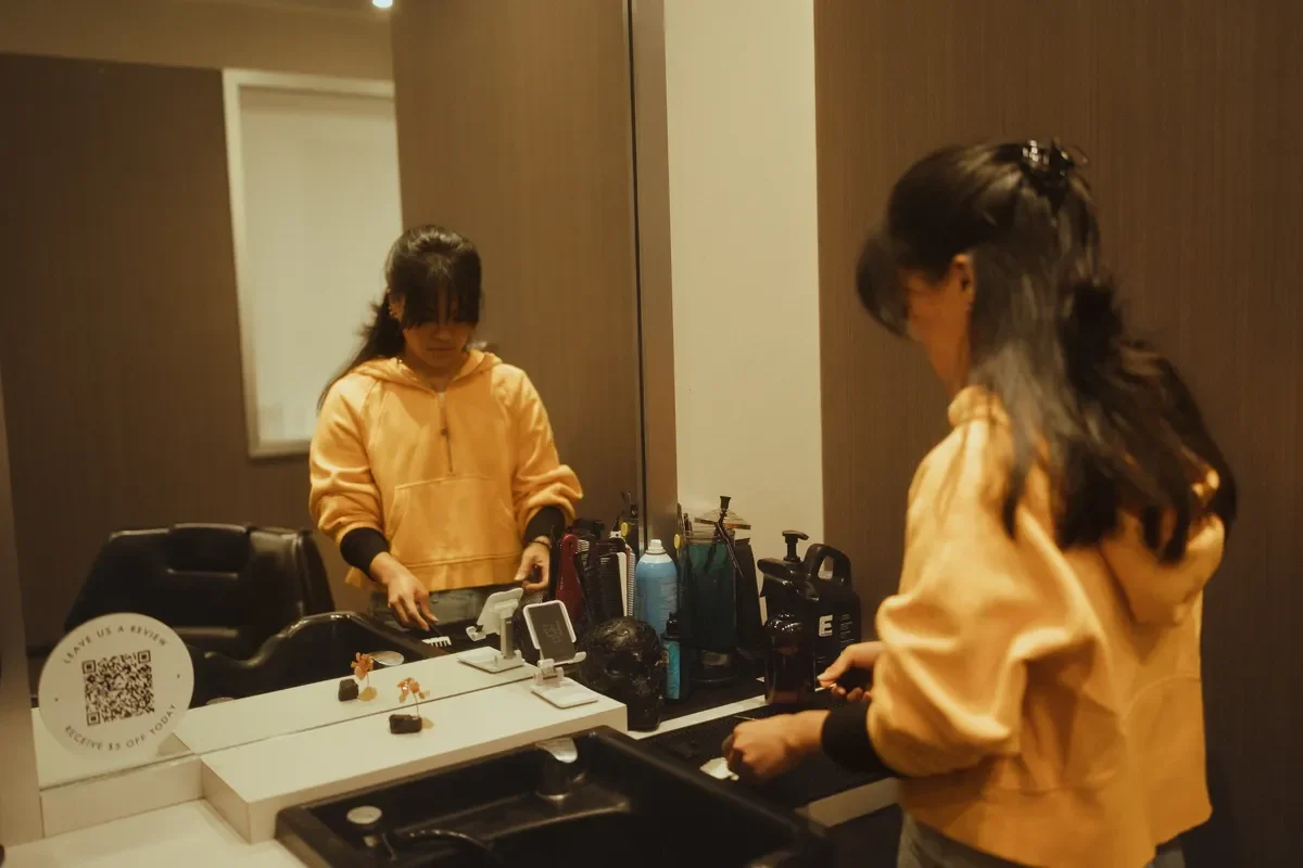 A young woman with long dark hair in a yellow hoodie looking into a mirror in a room with beige walls, with personal care items on the counter.