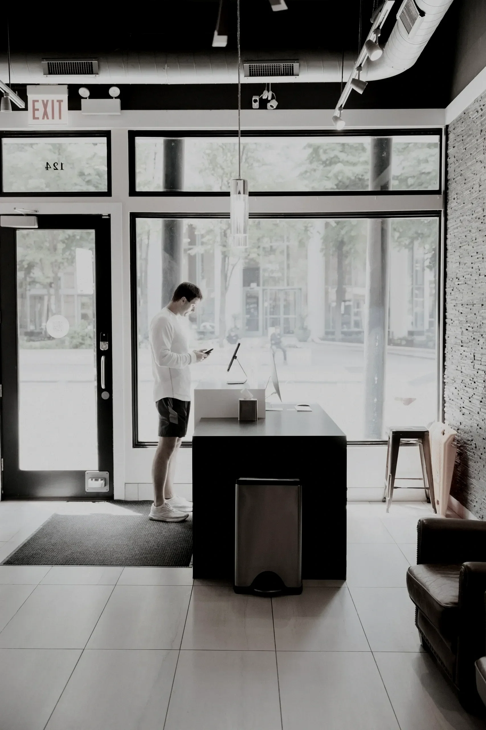 A man wearing a white long-sleeve shirt and shorts, standing inside a modern office or café, looking at his phone. The space has large glass windows and a door, with outdoor trees and buildings visible outside. The interior features minimalistic decor, a black table with a computer monitor, a bench on the right, an exit sign, and ceiling ventilation ducts.