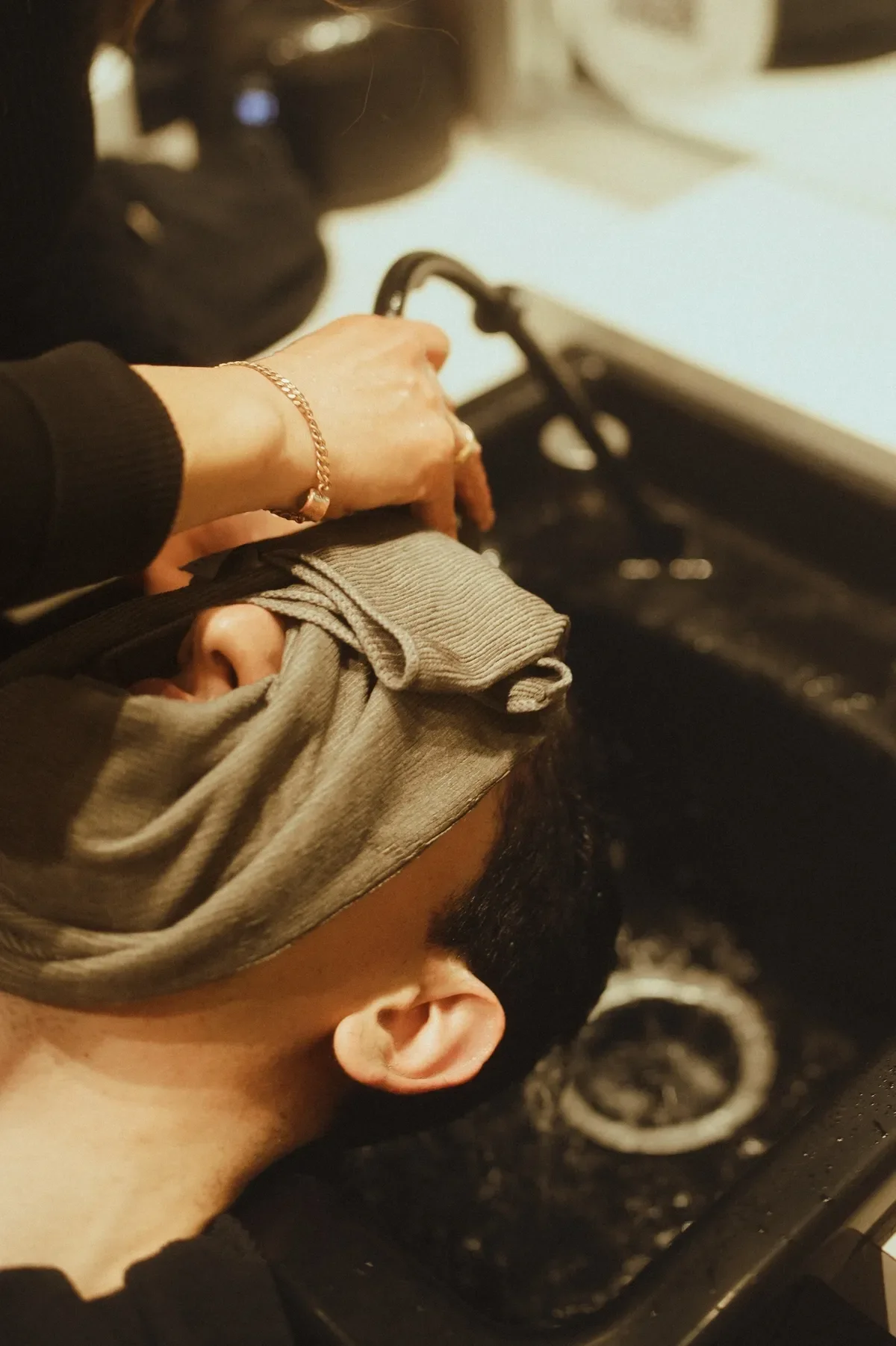 A person with short dark hair wearing a gray face mask and a beige hoodie, lying down with the head on a black surface. Someone else is holding the person's arm, which has a gold bracelet, over their face.