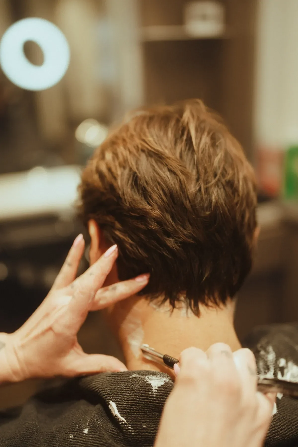 A person with short, layered, light brown hair is having their hair styled by a hairstylist. The stylist's hand is visible, holding a styling tool and touching the person's hair.