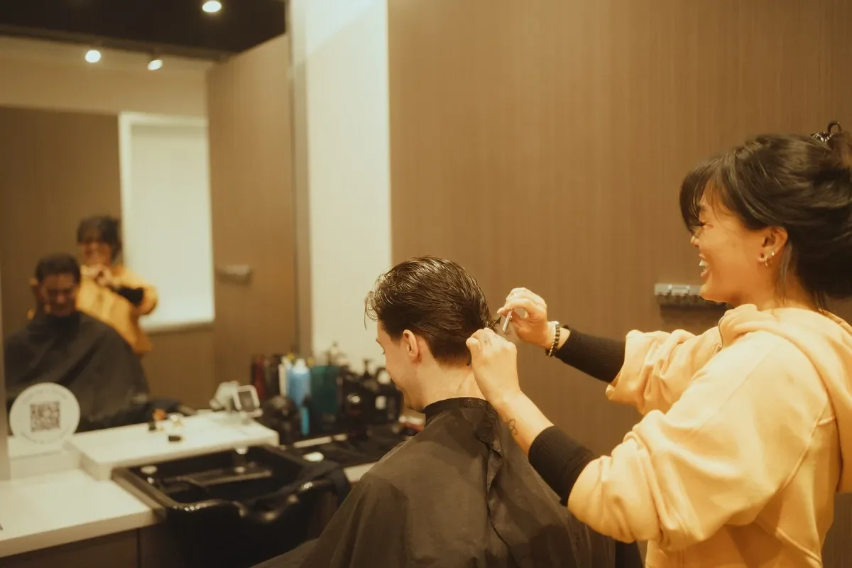 A hairstylist cutting a man's hair in a salon, with a mirror reflecting the scene, and another person in the background.