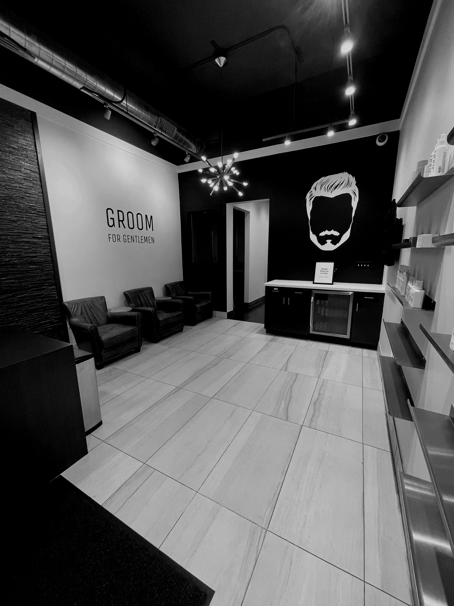 A waiting area in a barbershop with black chairs, a black and white wall art of a man with a beard and styled hair, and a sign that says 'GROOM FOR GENTLEMEN'. There is a contemporary chandelier lighting and a shelf with grooming products on the right side.