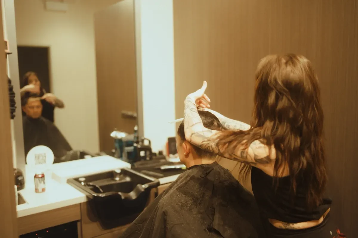 A hairdresser cuts and styles a man's hair in a salon with a large mirror, a counter with hairdressing tools, and a person visible in the reflection.