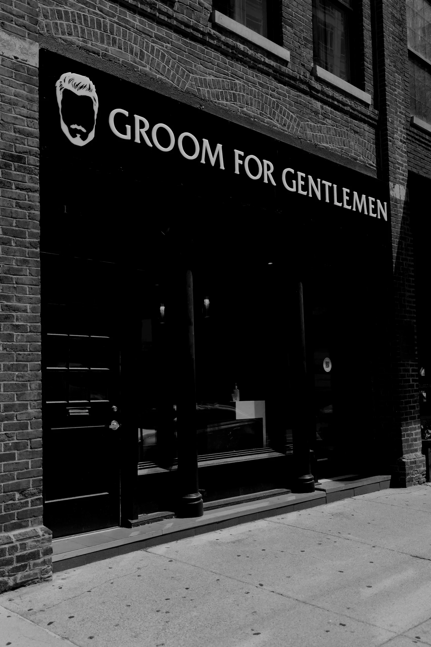 Storefront with a sign that reads 'Groom for Gentlemen' and an illustration of a man's face with a hair and beard style, outside on a city street.