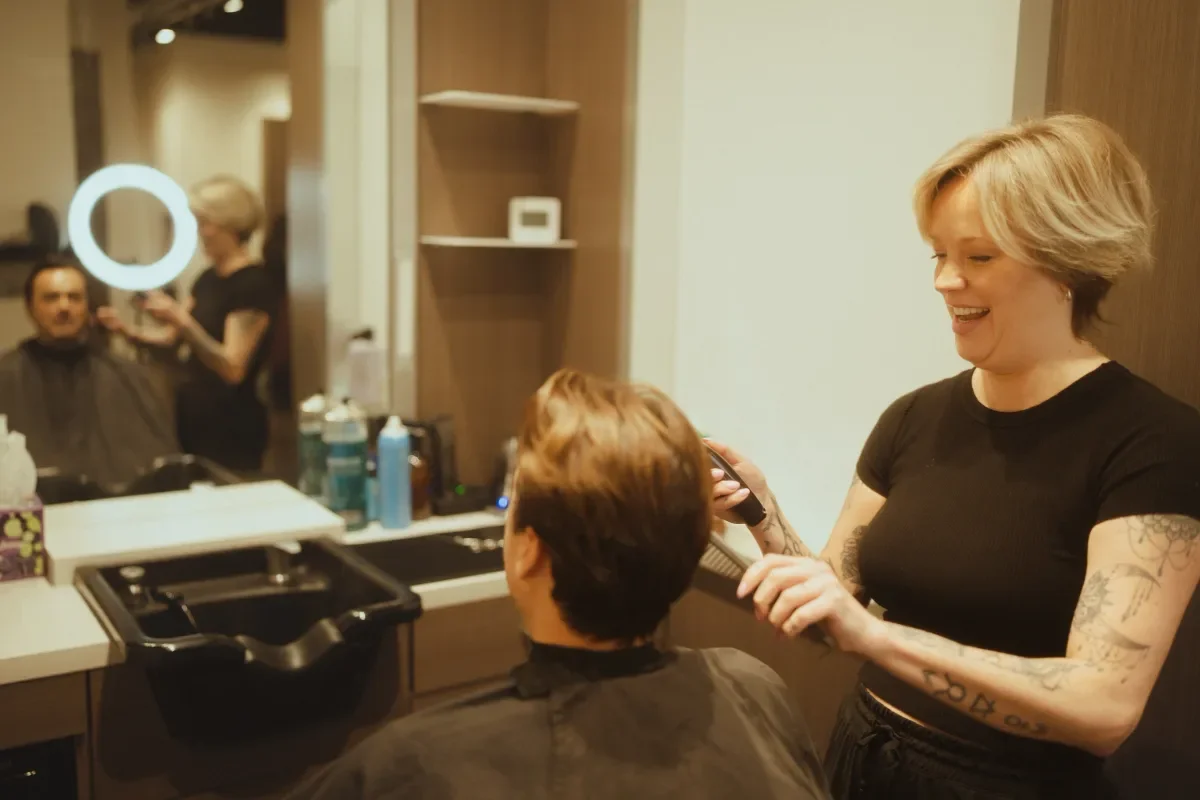 A woman is styling a man's hair in a salon. The woman has short blonde hair and tattoos on her arms, holding a styling tool and smiling. The man is seated in a salon chair, facing a mirror. There are hairstyling tools, products, and a ring light visi
