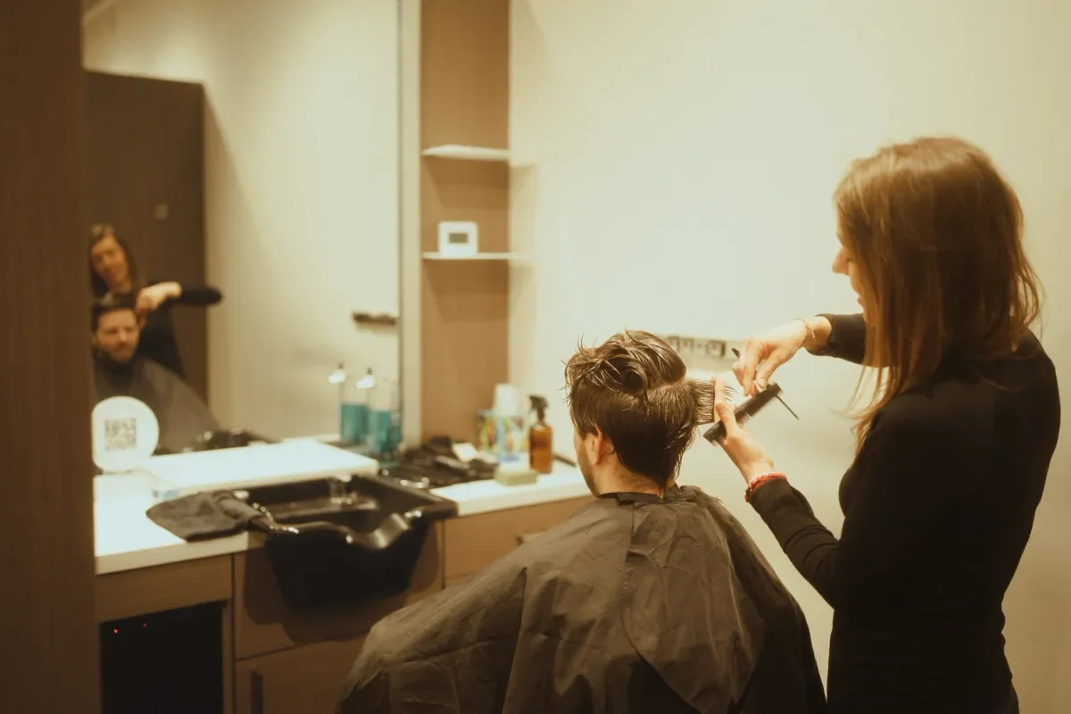 A hairstylist cutting a man's hair in a salon mirror, with another person visible in the reflection.