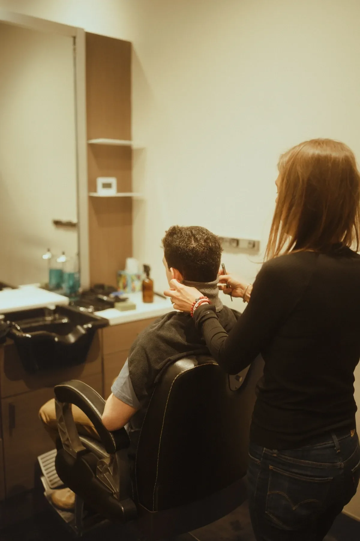 Woman styling a man's hair in a salon, with hairdressing tools and products on the counter nearby.