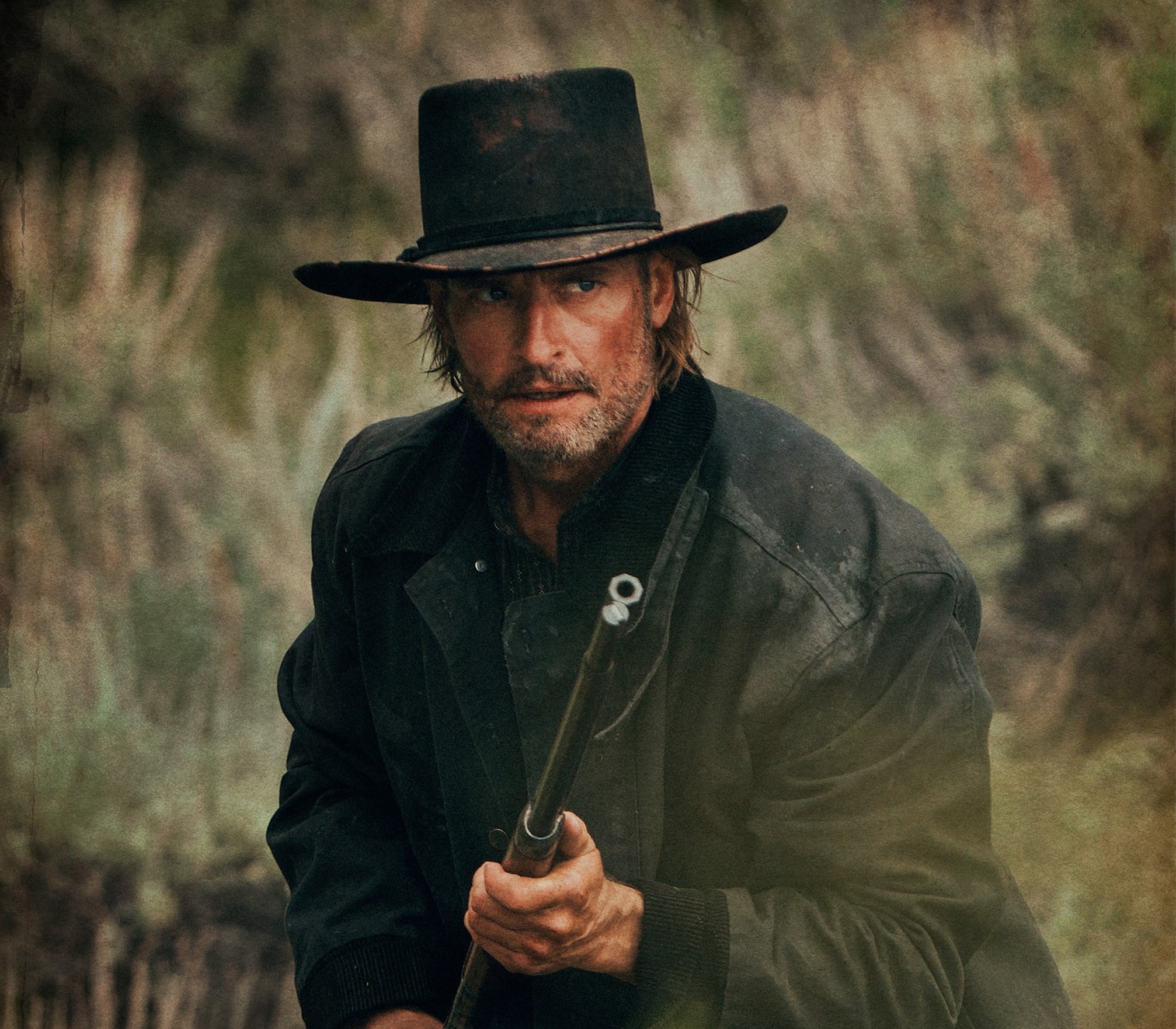Close-up of a man with long hair and beard, wearing a rugged black coat and a large, weathered wide-brimmed hat, holding an axe in his hand with a blurred natural outdoor background.