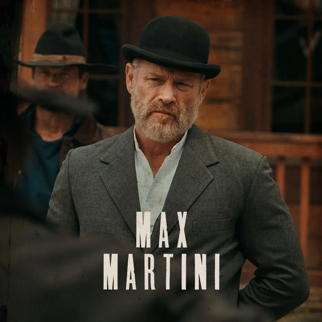 A man with a beard and mustache wearing a gray suit and a black bowler hat, standing outdoors with a wooden building in the background. The text 'Max Martini' is overlaid on the image.