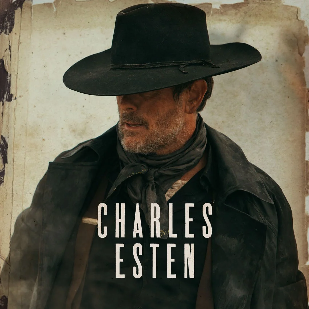 A man wearing a black wide-brimmed hat, a dark jacket, and a bandana around his neck, with a weathered background. Text on the image reads 'Charles Esten.'