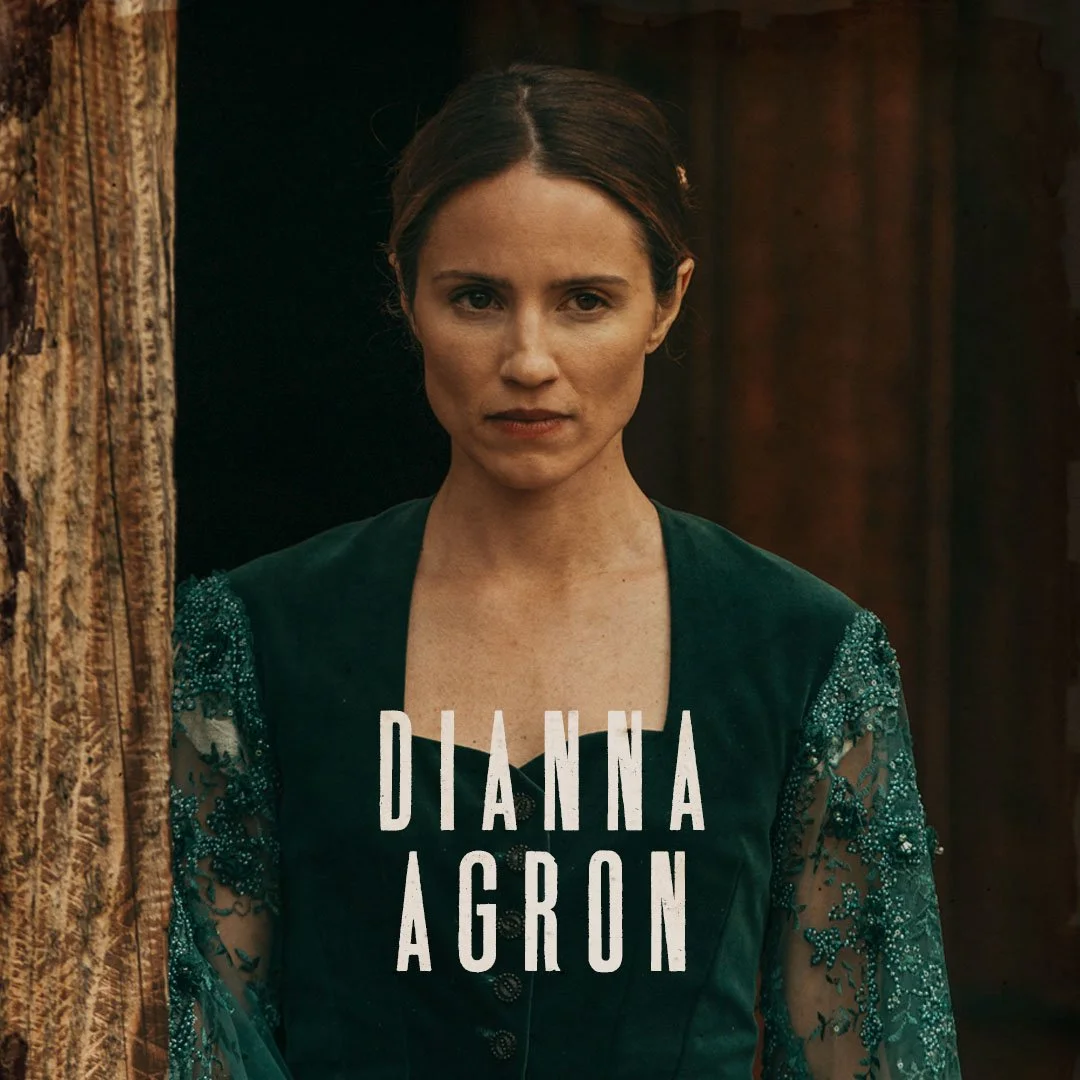 Close-up of a woman with brown hair and a serious expression, wearing a green dress with lace sleeves, standing next to a wooden beam.