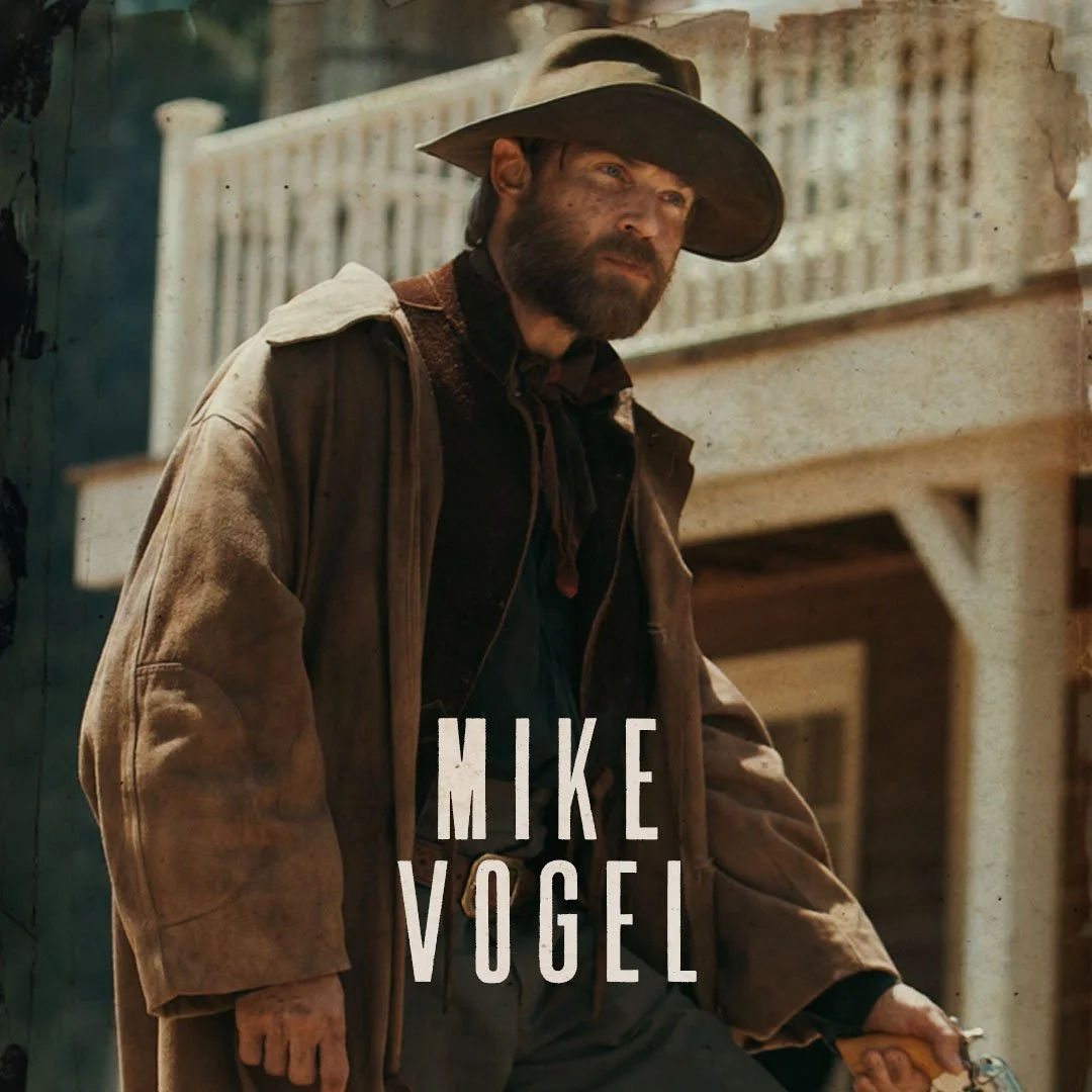 A man dressed as a cowboy with a large hat, beard, and dirty face, leaning on a fence on a western-style porch with buildings in the background. The text 'MIKE VOGEL' is overlaid at the bottom.