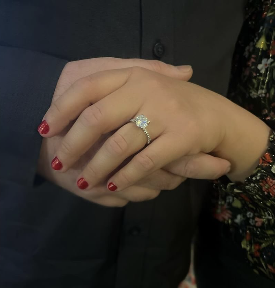 Close-up of two hands holding each other, one wearing an engagement ring with a large center diamond. The hands have red-painted fingernails.