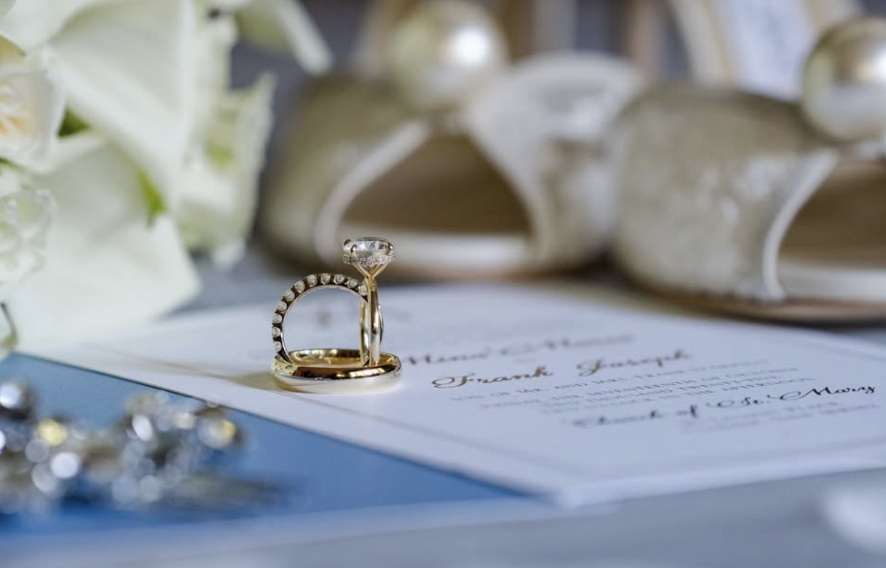 Close-up of wedding rings on a letter with a bouquet of white flowers and a pair of white shoes in the background.