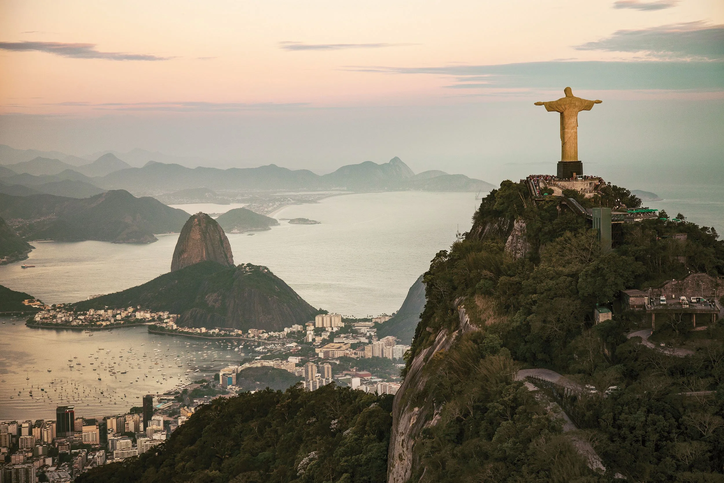 View of Rio de Janeiro with Christ the Redeemer statue on a hill at sunrise or sunset, overlooking the city, mountains, and water.