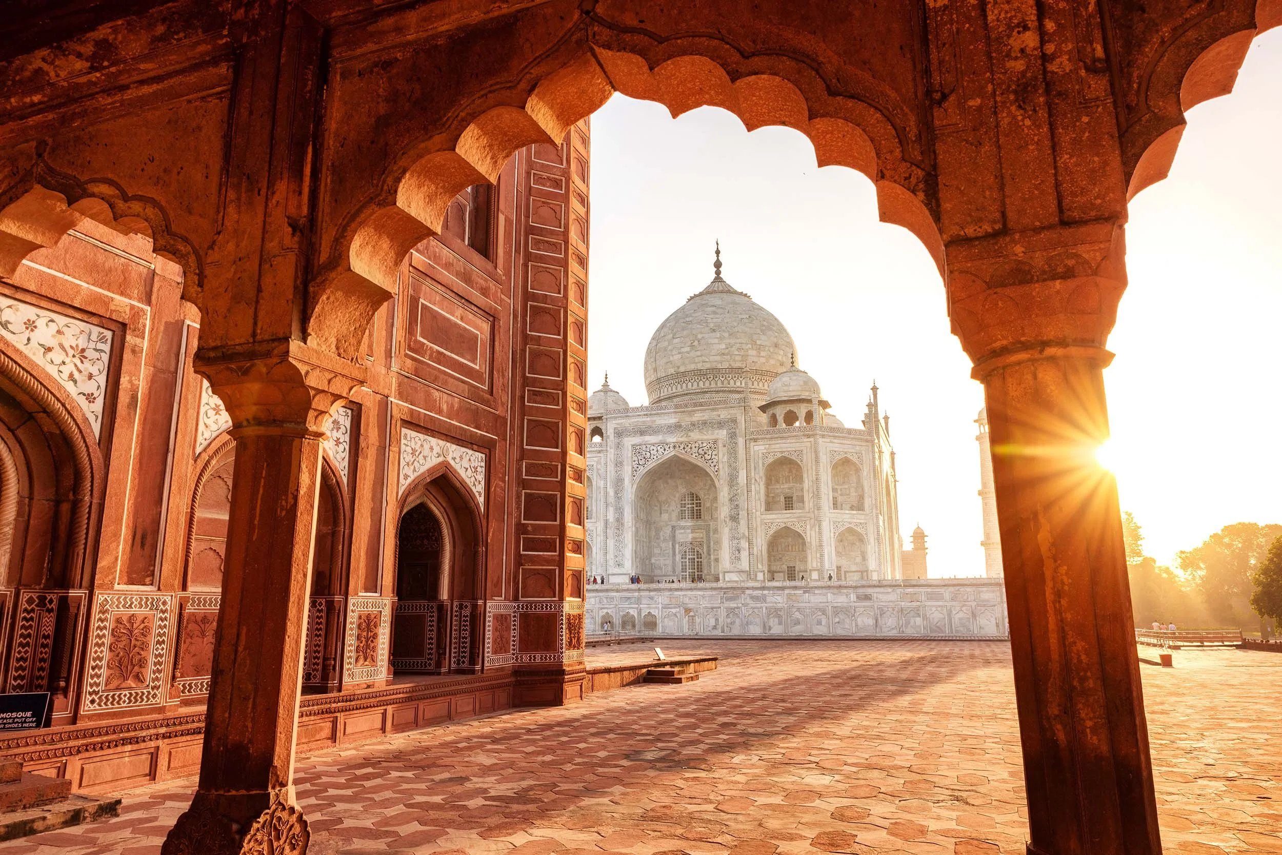The image shows the Taj Mahal in India seen through an ornate archway with reddish sandstone and intricate carvings. The white marble mausoleum is illuminated by the golden sunlight of sunset.