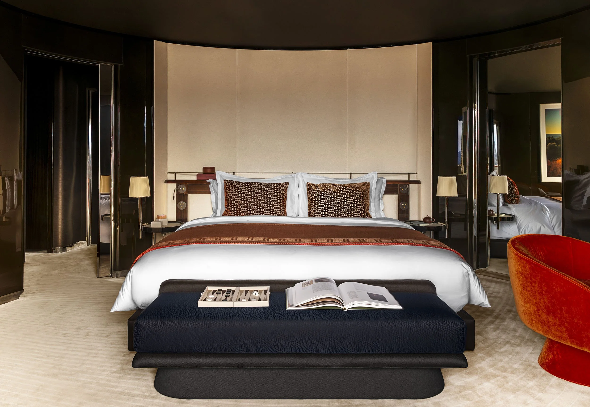 The Zephyr Suite on board The Orient Express Corinthian, with a large bed with white linens and decorative pillows, a black bench at the foot of the bed with a book and a chess set, two bedside lamps,.