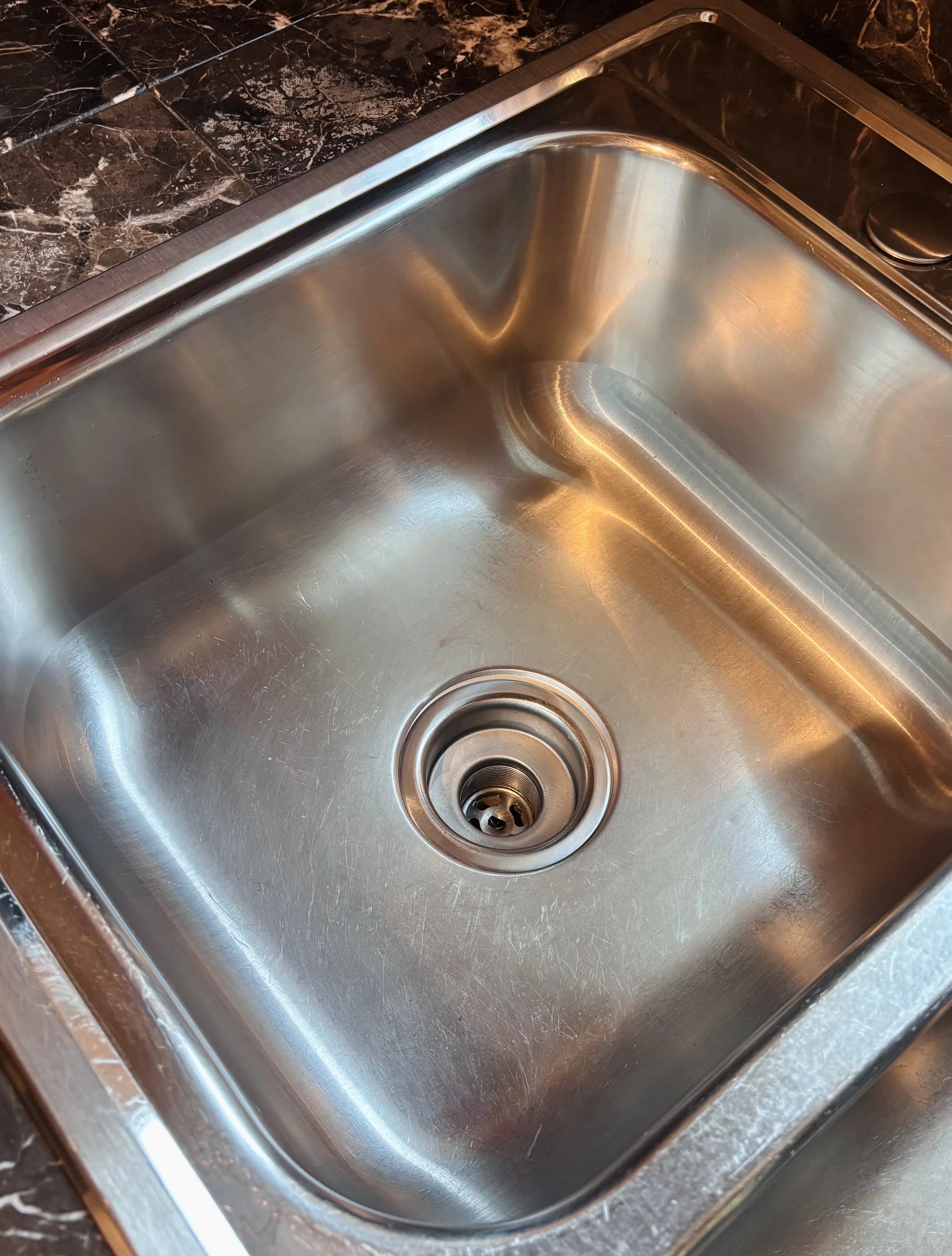 Stainless steel kitchen sink with drain, set into a dark marble countertop.