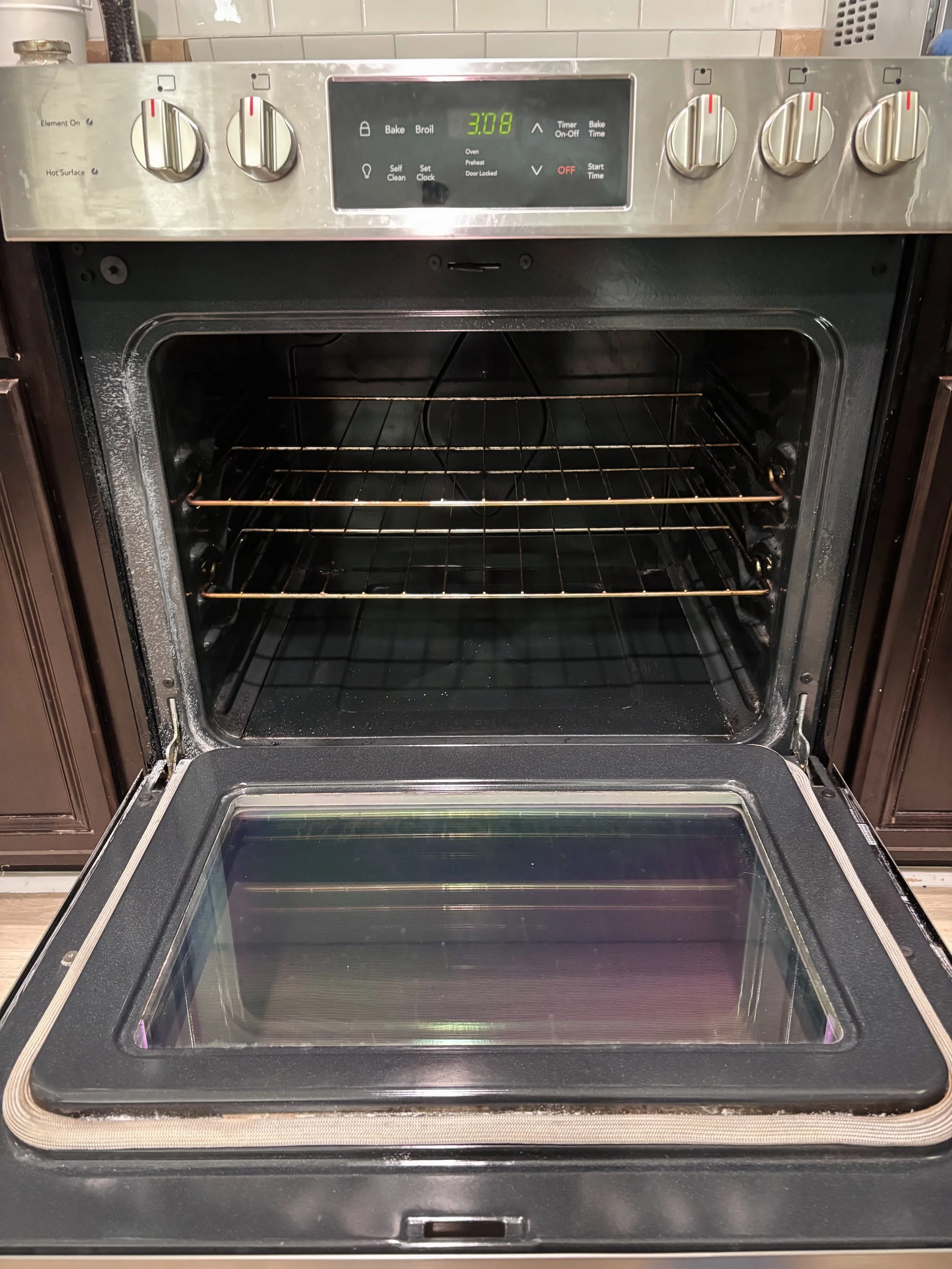An open electric oven with a digital display showing 3:08. The oven has four dials on the top panel, a glass oven door, and interior racks.