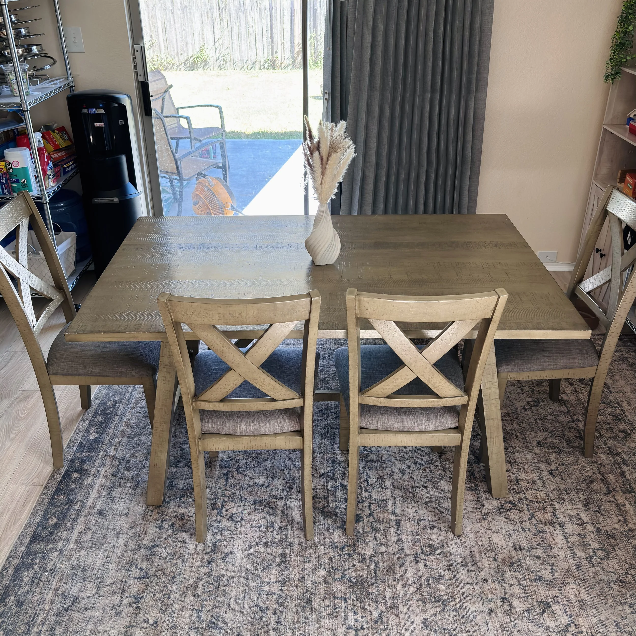 A dining table with a vase containing dried pampas grass, surrounded by wooden chairs with padded seats, on a patterned rug in a room with a sliding glass door, curtains, and shelves with various household items.