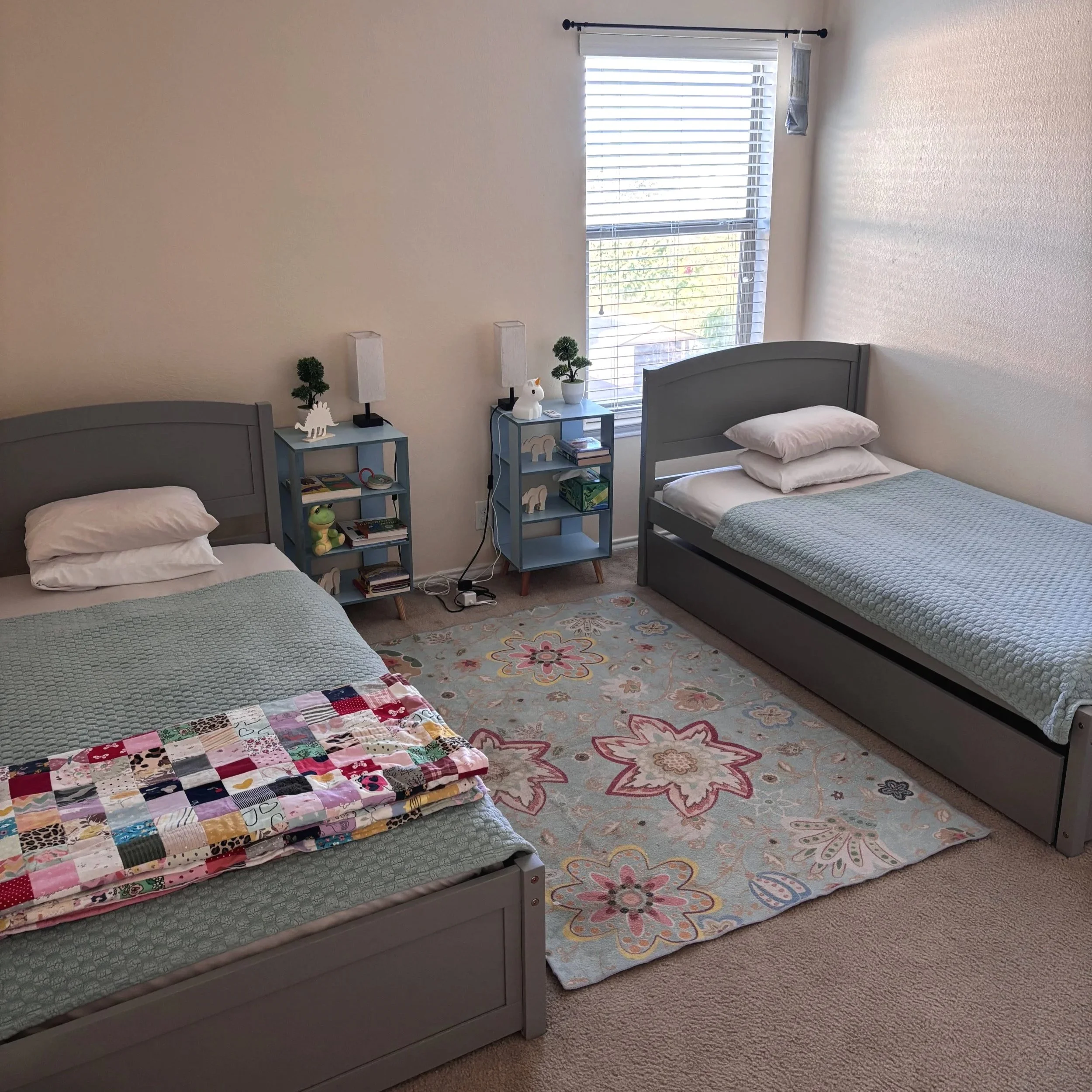 A bedroom with two twin beds with gray headboards and matching light blue quilts. Each bed has white pillows. Between the beds, there are two small blue nightstands, each decorated with a small potted plant, a white unicorn, and a small wooden animal figure. One nightstand has books and a remote. There are two white lamps on the wall above each bed. A multicolored quilt is folded on the left bed. The room has a large window with blinds, and a floral area rug is on the beige carpet.