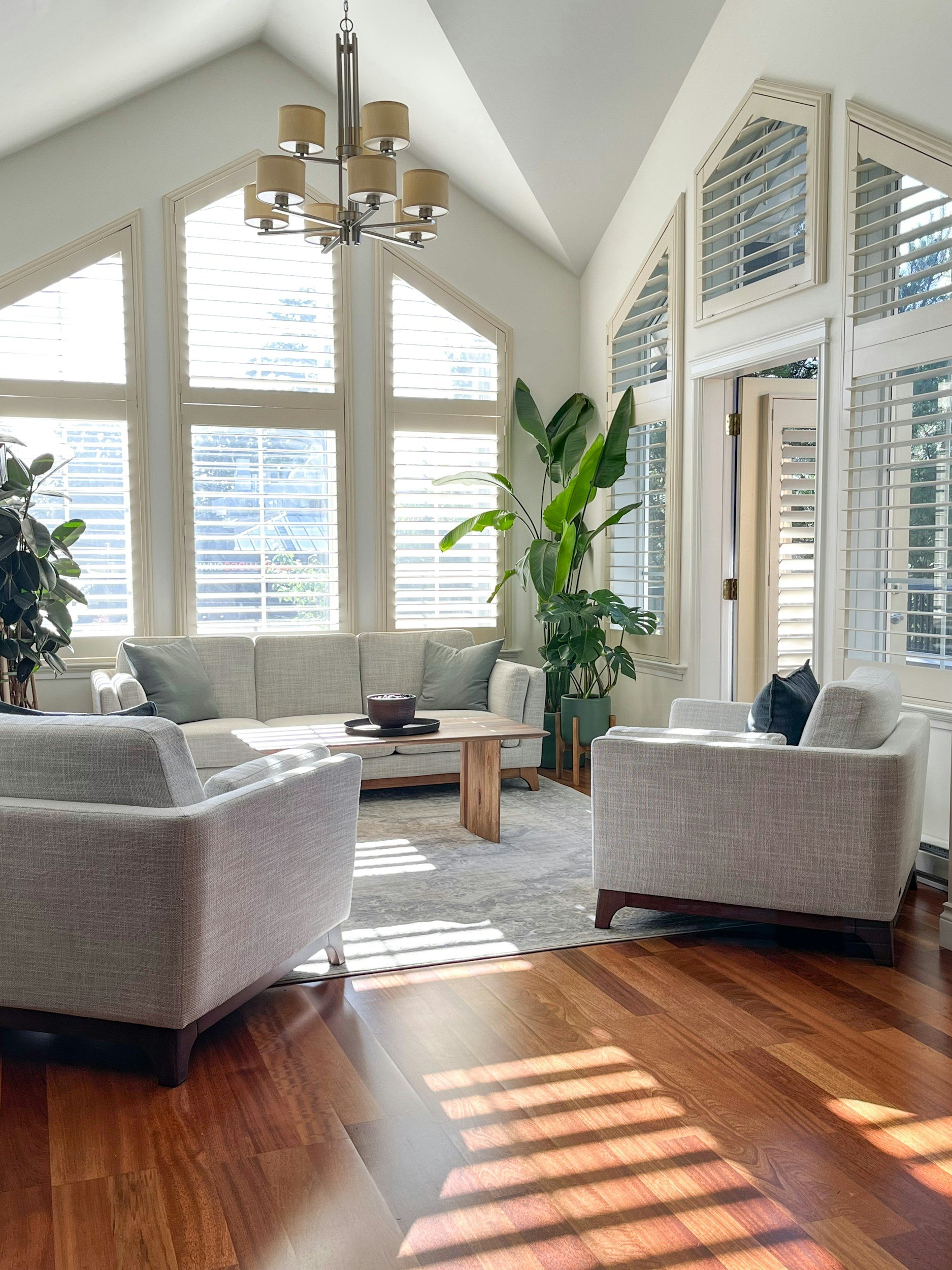 A living room with white sofas, beige cushions, a wooden coffee table with a bowl, large windows with white shutters, a tall green plant, a chandelier, and hardwood floors.