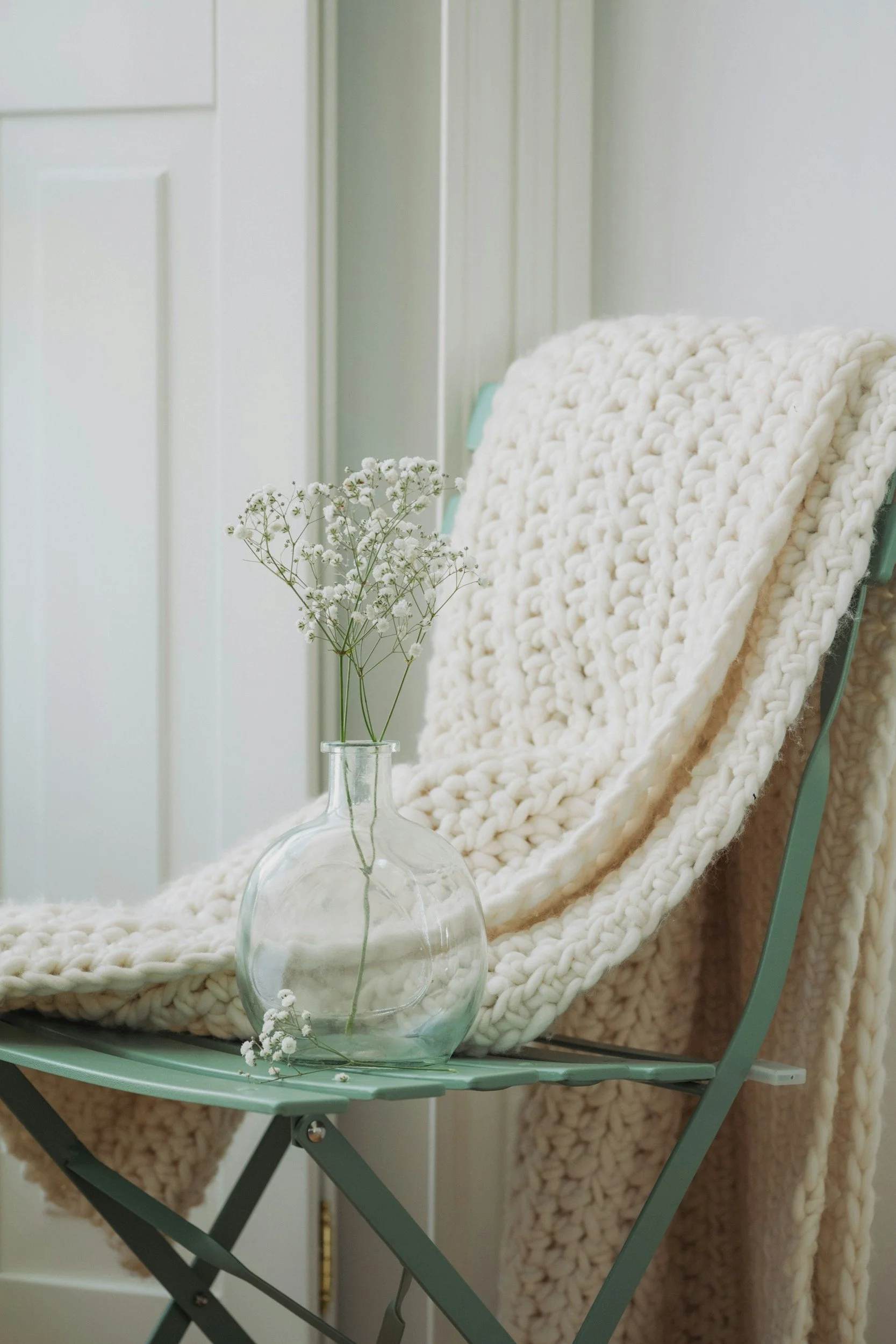 A glass vase with small white flowers placed on a pastel green folding table. In the background, there is a white, chunky knit blanket draped over a chair or bench, with a light-colored wall and a door.
