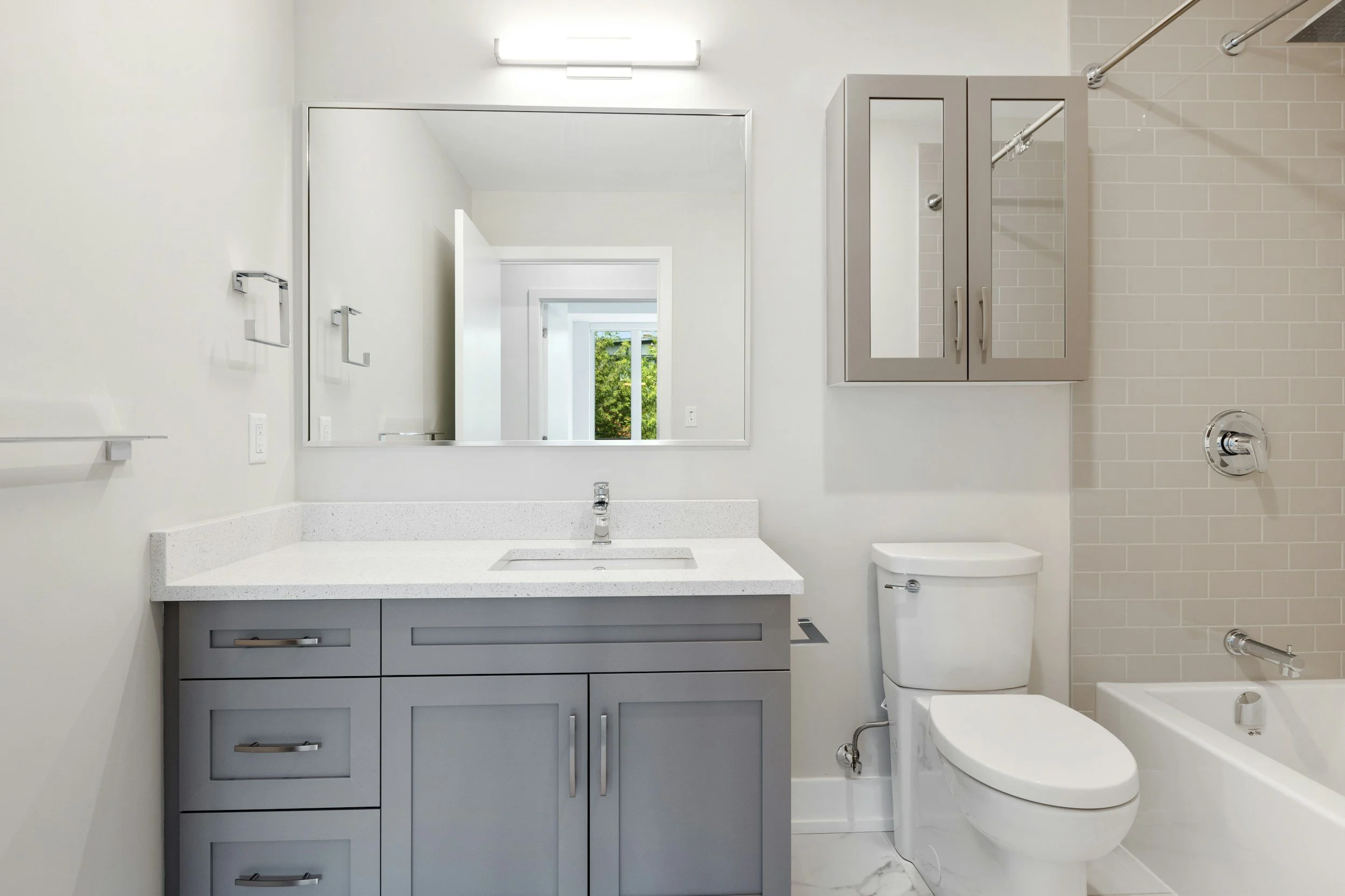A modern bathroom with a mirror, sink, gray cabinet, toilet, and shower area with glass and tile walls.