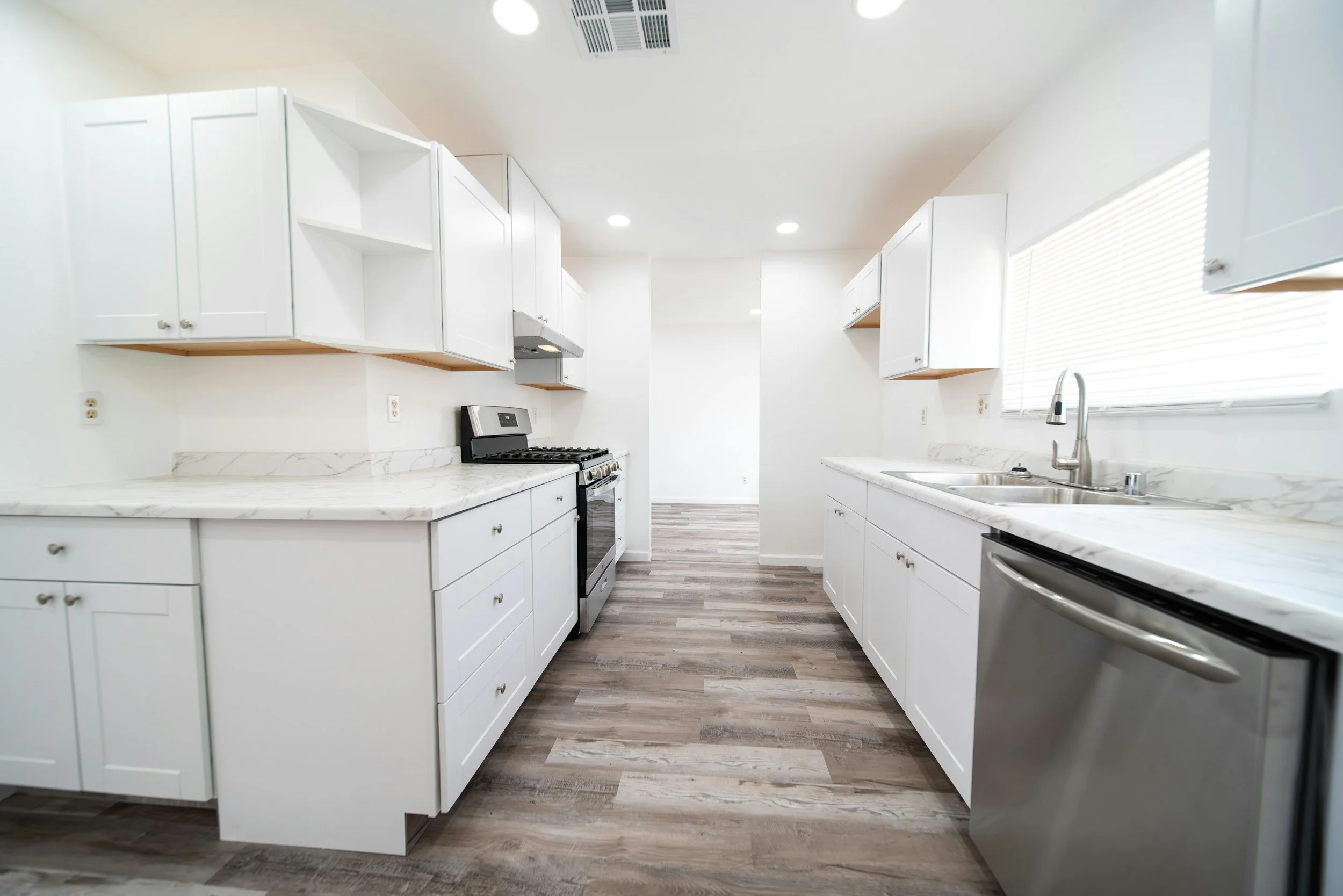 Modern kitchen with white cabinets, marble countertops, stainless steel appliances, a double sink, and wood-look flooring, illuminated by ceiling lights and natural light through window blinds.