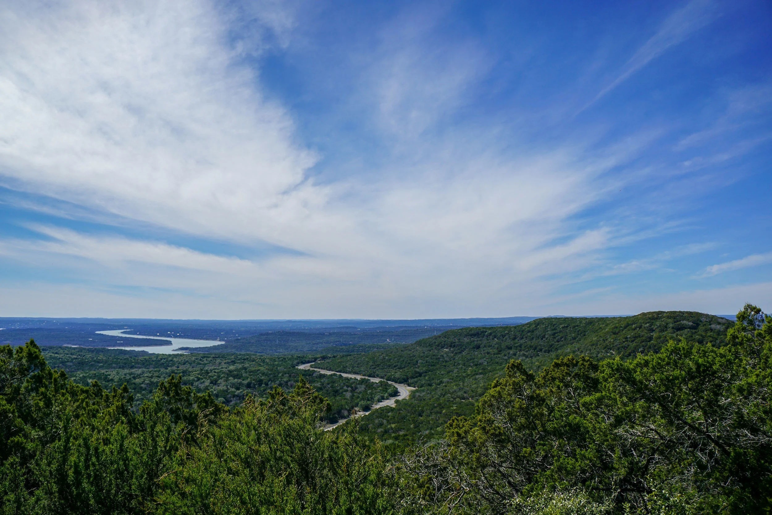 View of lush green hills, a winding road, and a river in the distance under a partly cloudy blue sky.