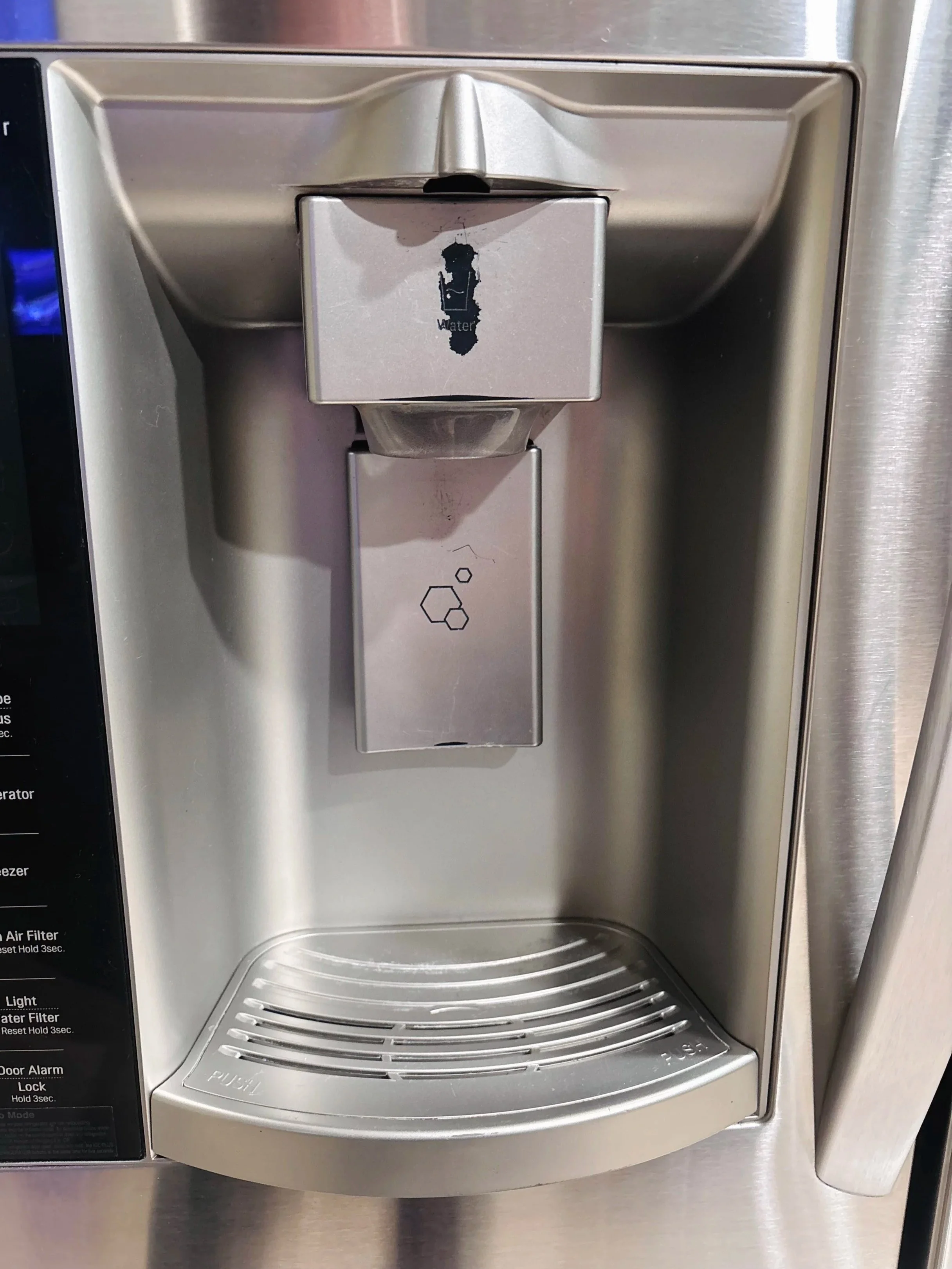 A stainless steel water dispenser with a black and white water droplet icon and a textured drip tray at the bottom.