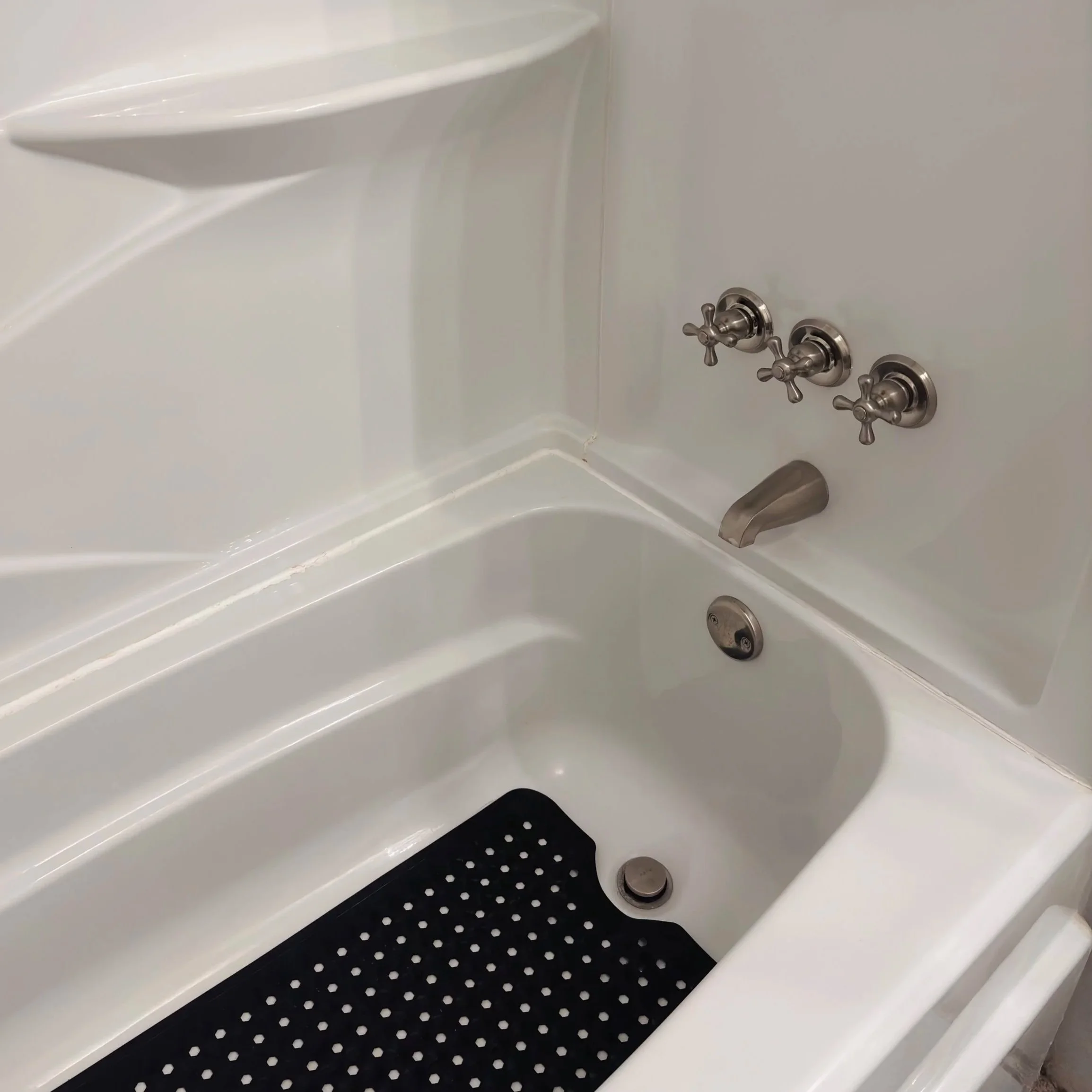 White shower or bathtub with three silver faucets and a black anti-slip mat with white dots.