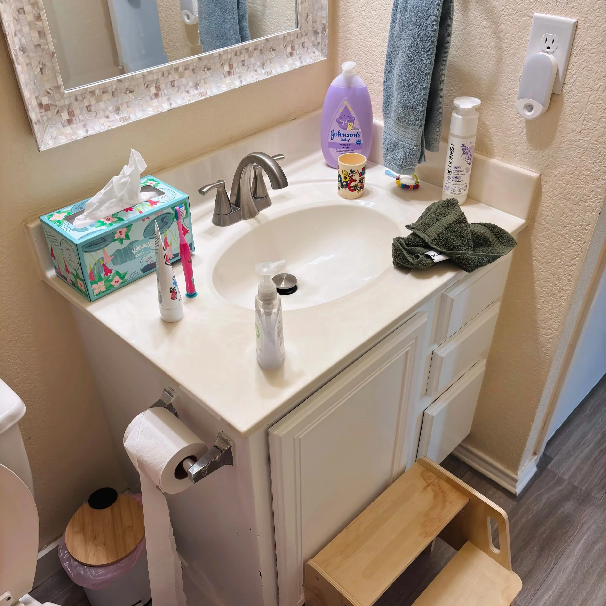 Bathroom sink with various items including a tissue box, toothbrushes, mouthwash, lotion, hairbrush, hand soap, towel, and a wooden shelf below the sink. Mirror with decorative frame hangs above the sink.