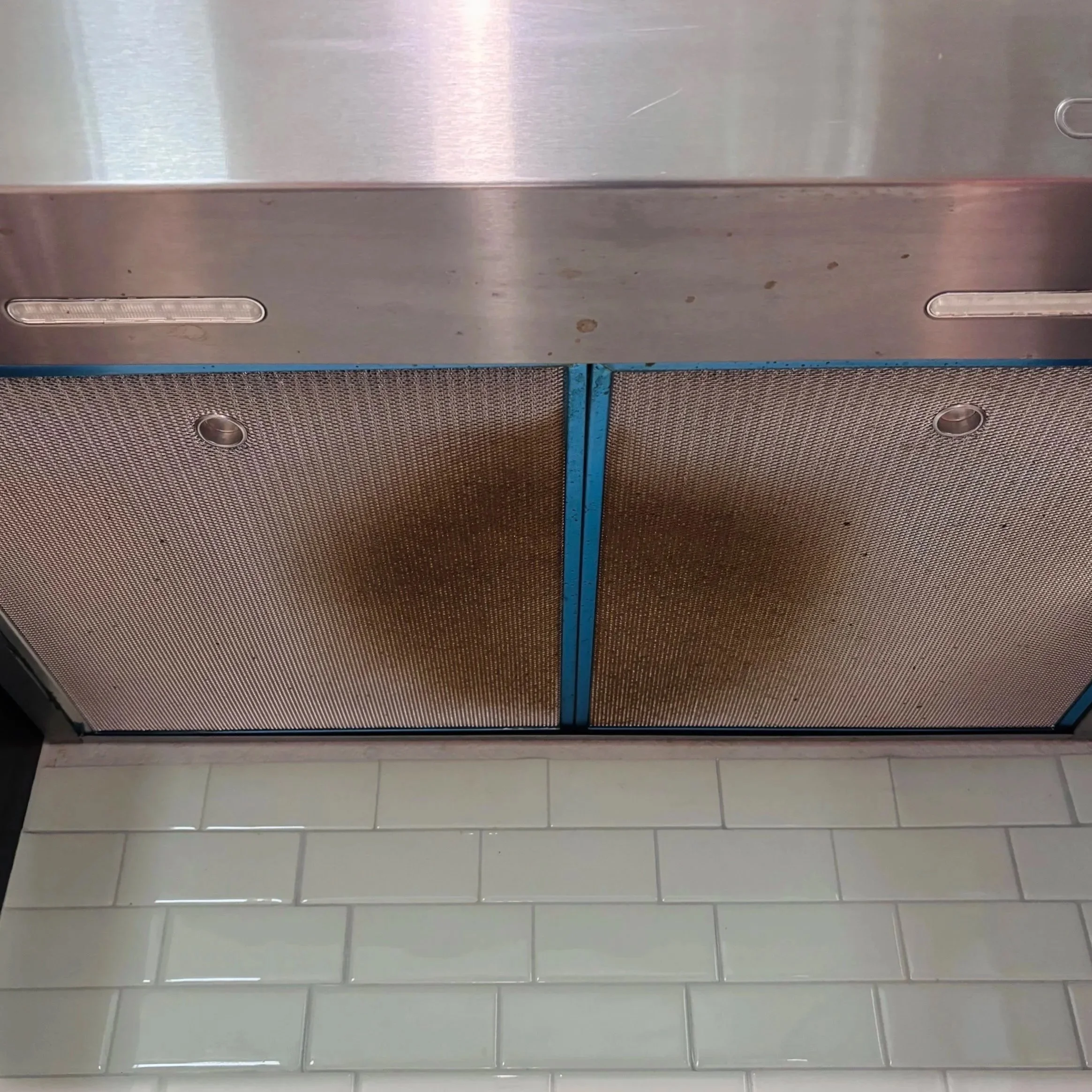 A metal mesh-covered speaker or air vent on a tiled wall with a row of white tiles and a tiled floor below.