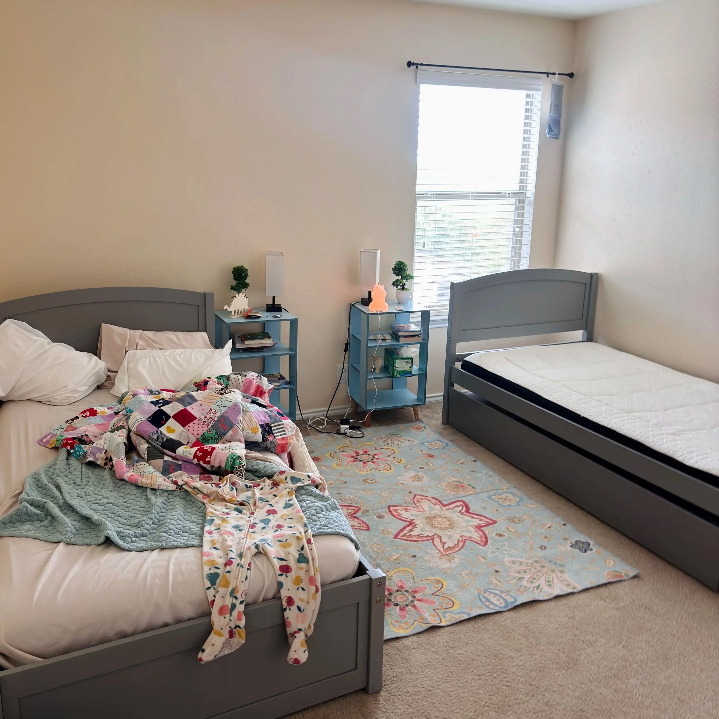 A bedroom with a large window covered by white blinds, two twin beds with gray headboards, one bed unmade with a colorful quilt. There are two blue nightstands, decorations, and a lamp, with a patterned area rug and beige carpet.