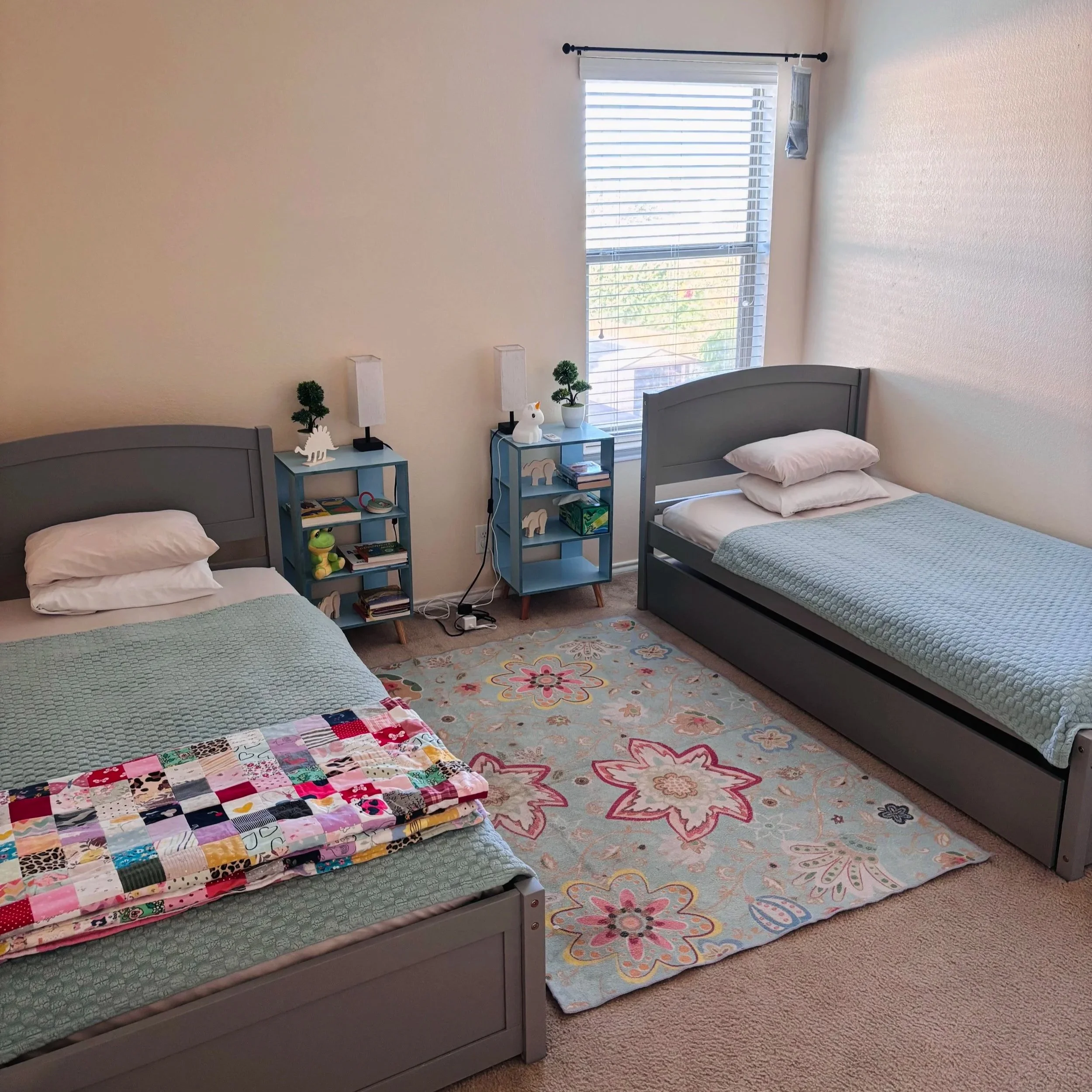 A bedroom with two beds, each with a light blue quilt and white pillows. Between the beds are two small blue nightstands holding decorative items, including a small potted plant and a white bunny sculpture. There is a window with white blinds above a