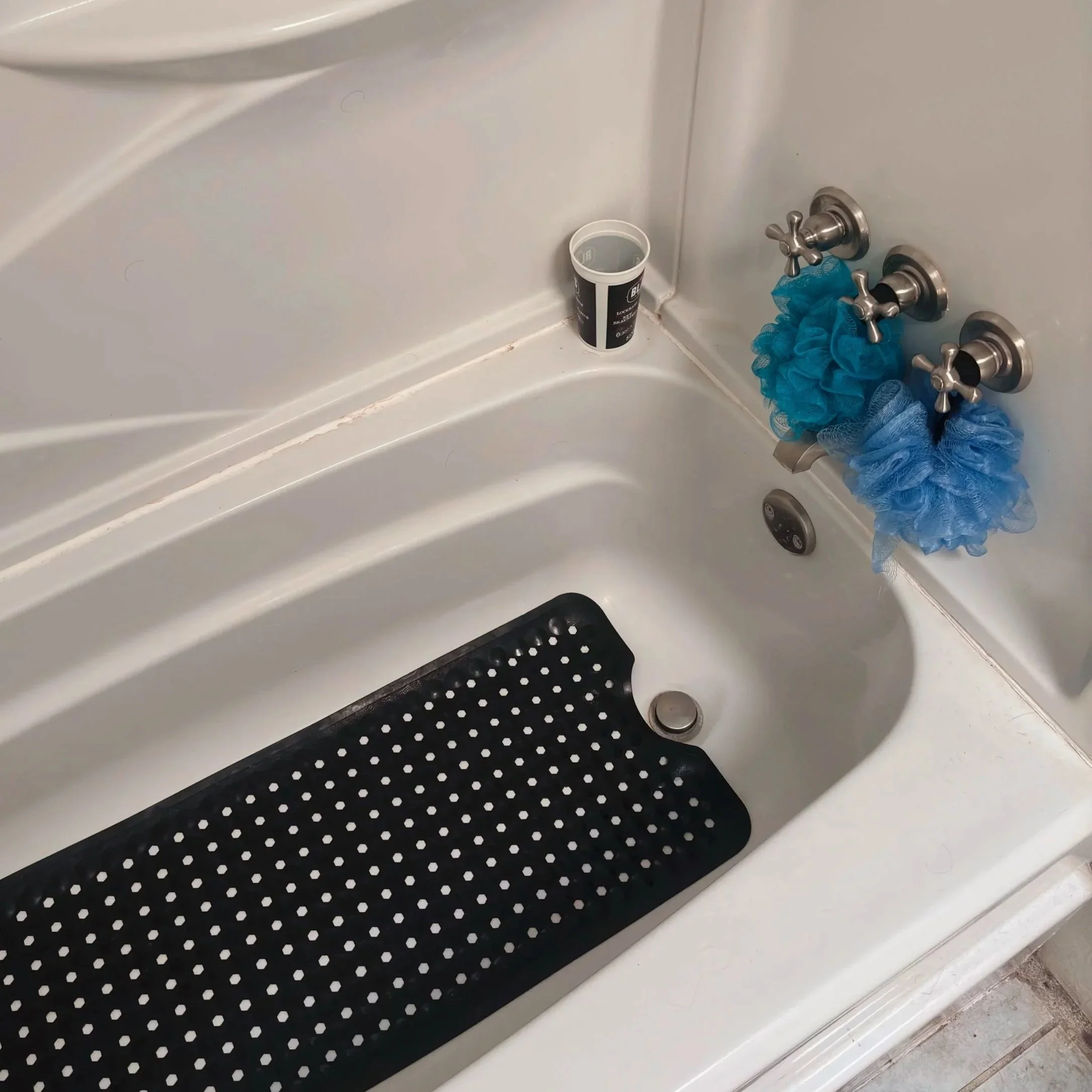 A bathtub with a black rubber mat at the bottom, two silver faucet handles with hub caps, and a bucket with a black and white label next to the faucet. A blue shower sponge hangs from the faucet handles.