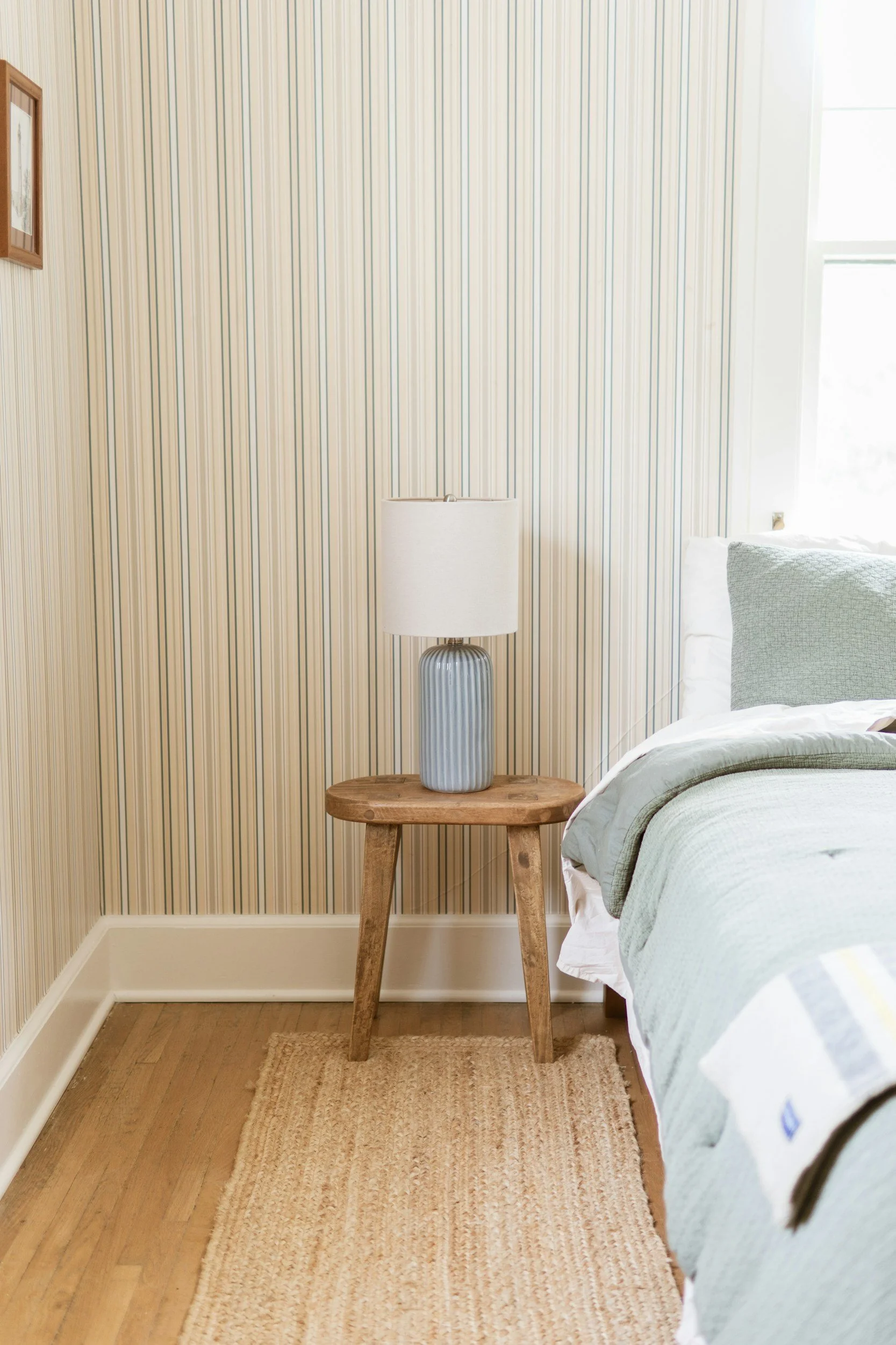 A bedroom corner with striped wallpaper, a wooden side table, a white table lamp, part of a bed with green and white bedding, a beige rug, and a window on the right side.