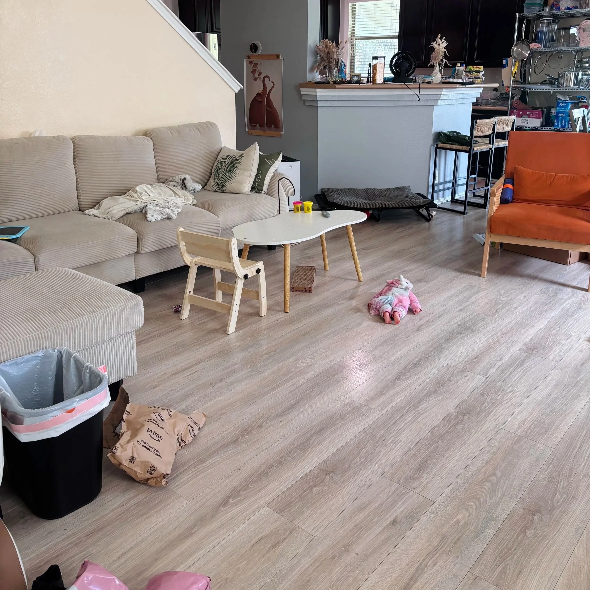 A living room with a beige couch, pillows, a small child's chair, a table with toys and remote, a pet dog sleeping on the couch, a trash can, and various household items and kitchen in the background.