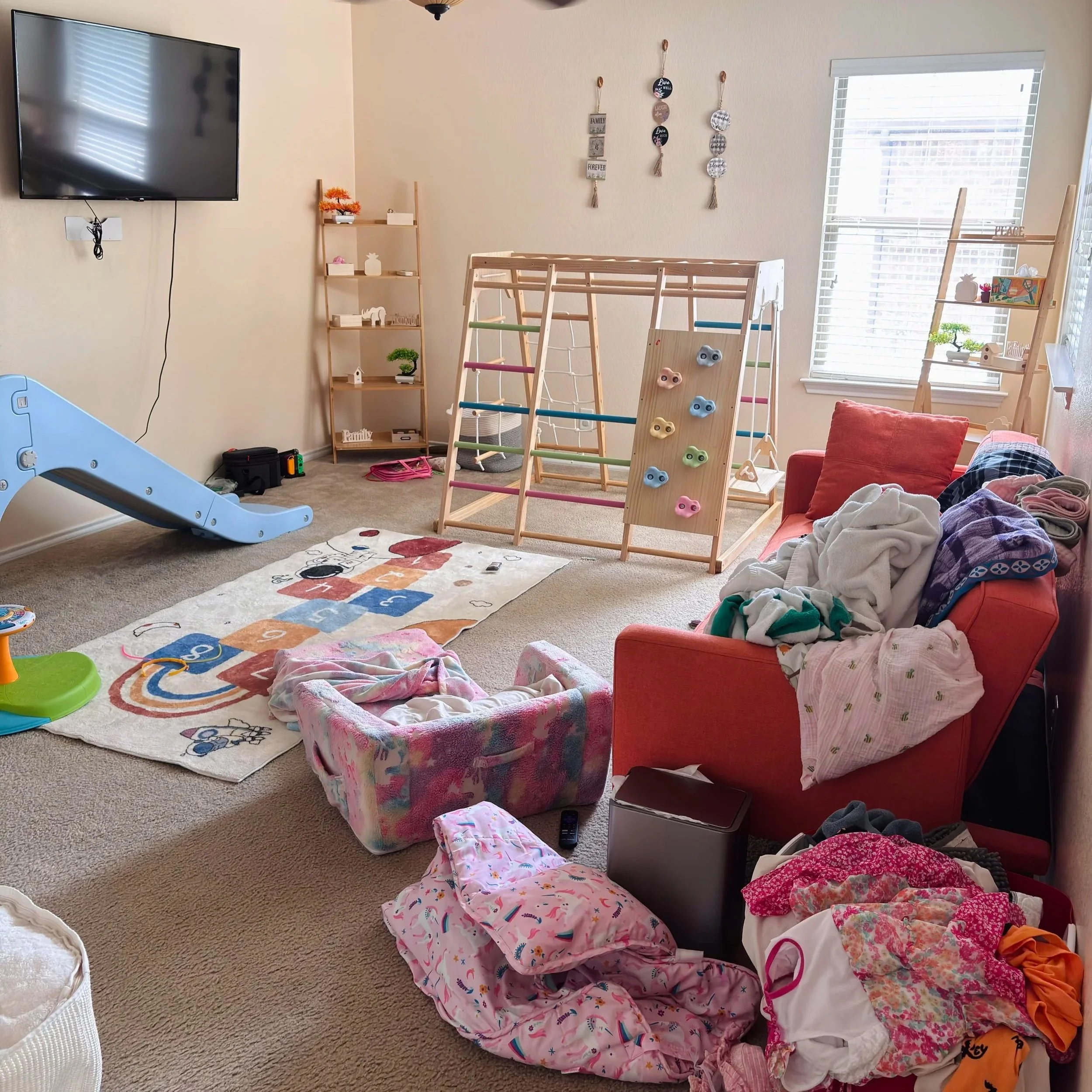 A cluttered living room with clothes and laundry on the floor, a small toddler slide, a colorful rug, a red couch with pillows, and a wooden shelving unit with decorative items and toys. The room has a window with blinds, and a large wall-mounted TV.