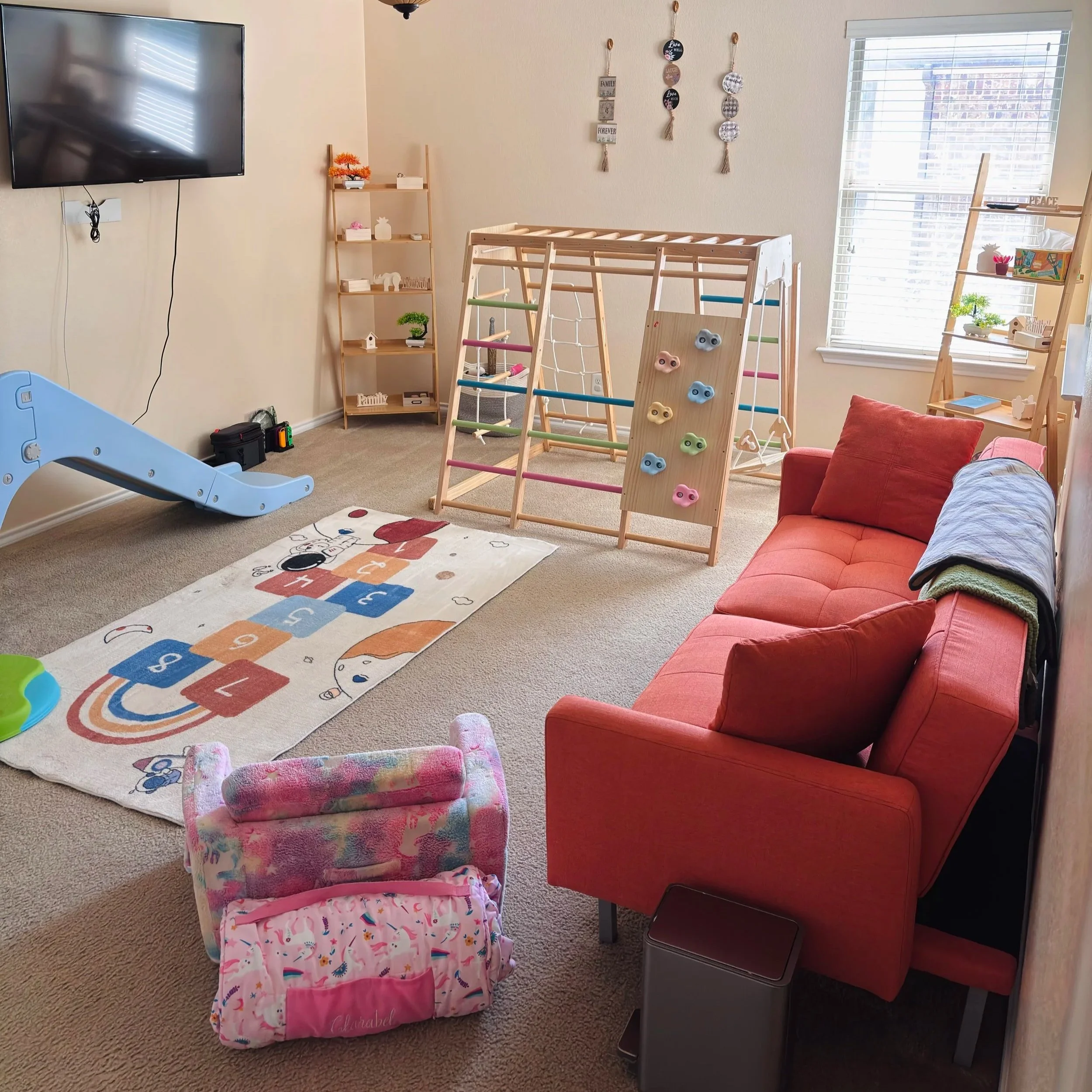 A bedroom with a children's area, featuring a blue slide, colorful climbing wall, a pink toy box, a red couch, a wooden ladder shelf with decorative items, a small white bench, and wall decorations.